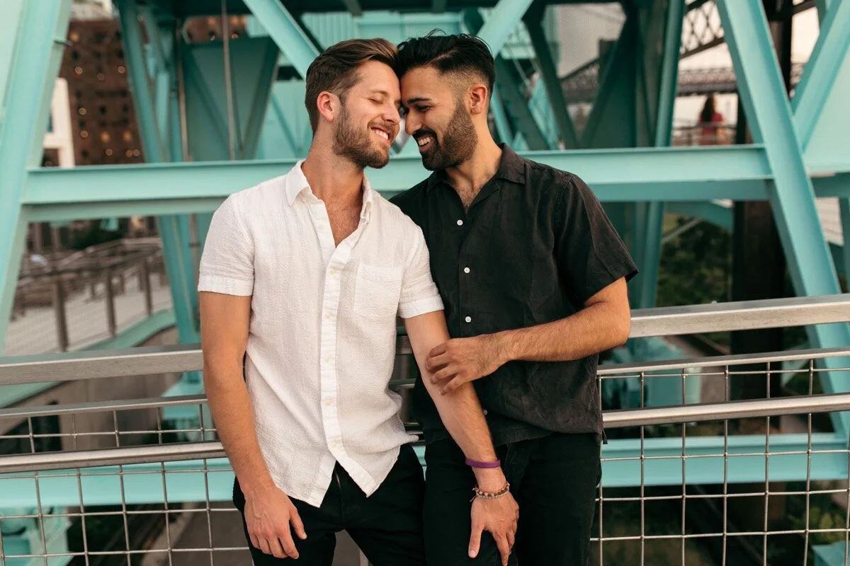 Joe and Rohan snuggle up together on walkway by bridge during couples photo session in Domino Park Brooklyn Bailey Q Photo