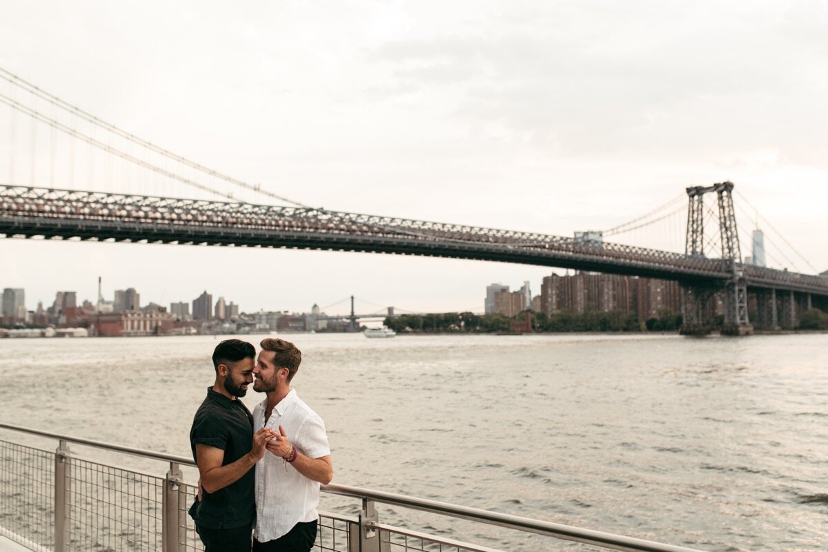 Joe and Rohan dance together by waterfront with bridge in background during couples photo session in Domino Park Brooklyn Bailey Q Photo
