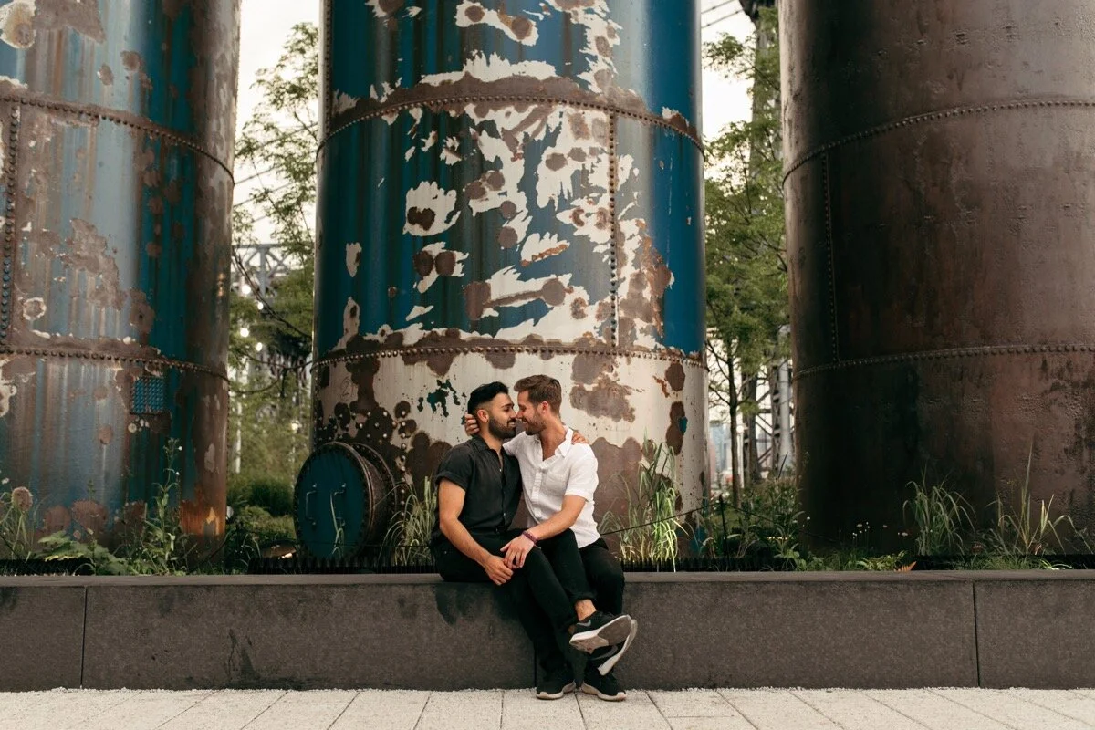 Joe and Rohan snuggle up together in front of industrial silos during couples photo session in Domino Park Brooklyn New York Bailey Q Photo