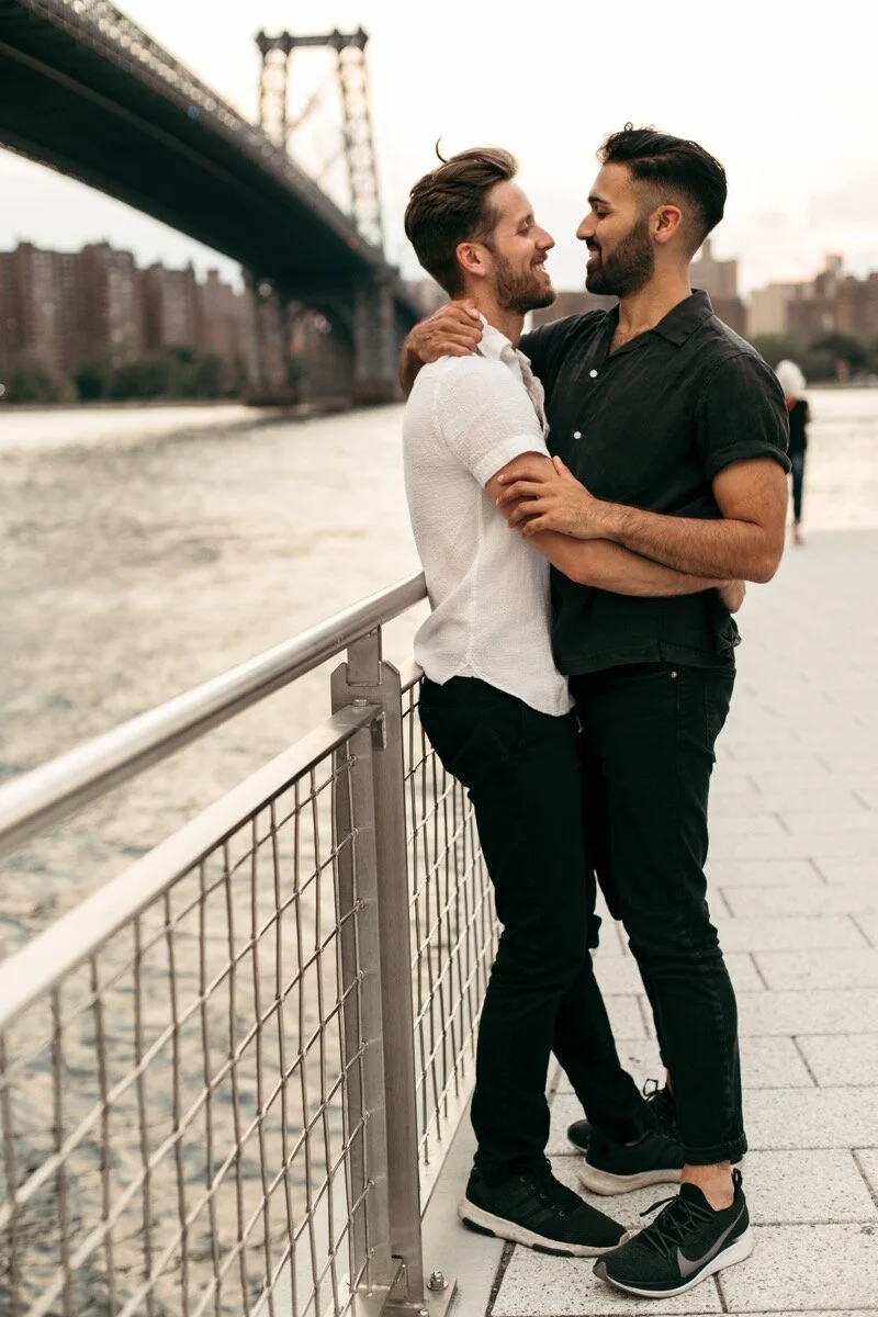 Joe and Rohan snuggle up together along the waterfront during couples photo session in Domino Park Brooklyn New York Bailey Q Photo