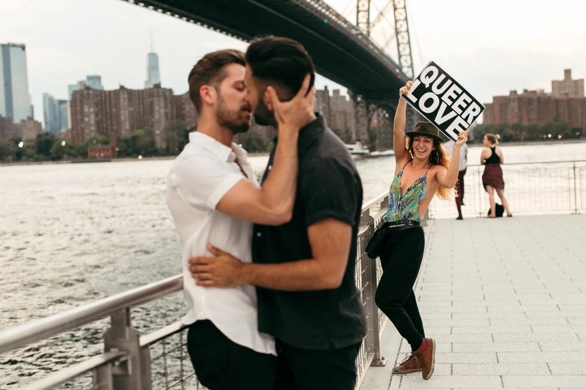Joe and Rohan kiss along the waterfront while passerby holds sign that says “Queer Love” during couples photo session in Domino Park Brooklyn New York Bailey Q Photo