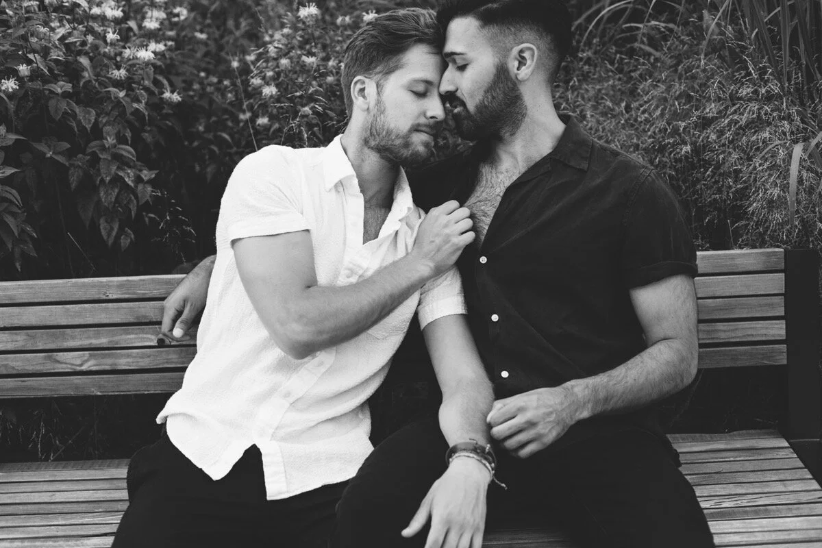 Joe and Rohan snuggle up together on wooden bench during couples photo session in Domino Park Brooklyn Bailey Q Photo