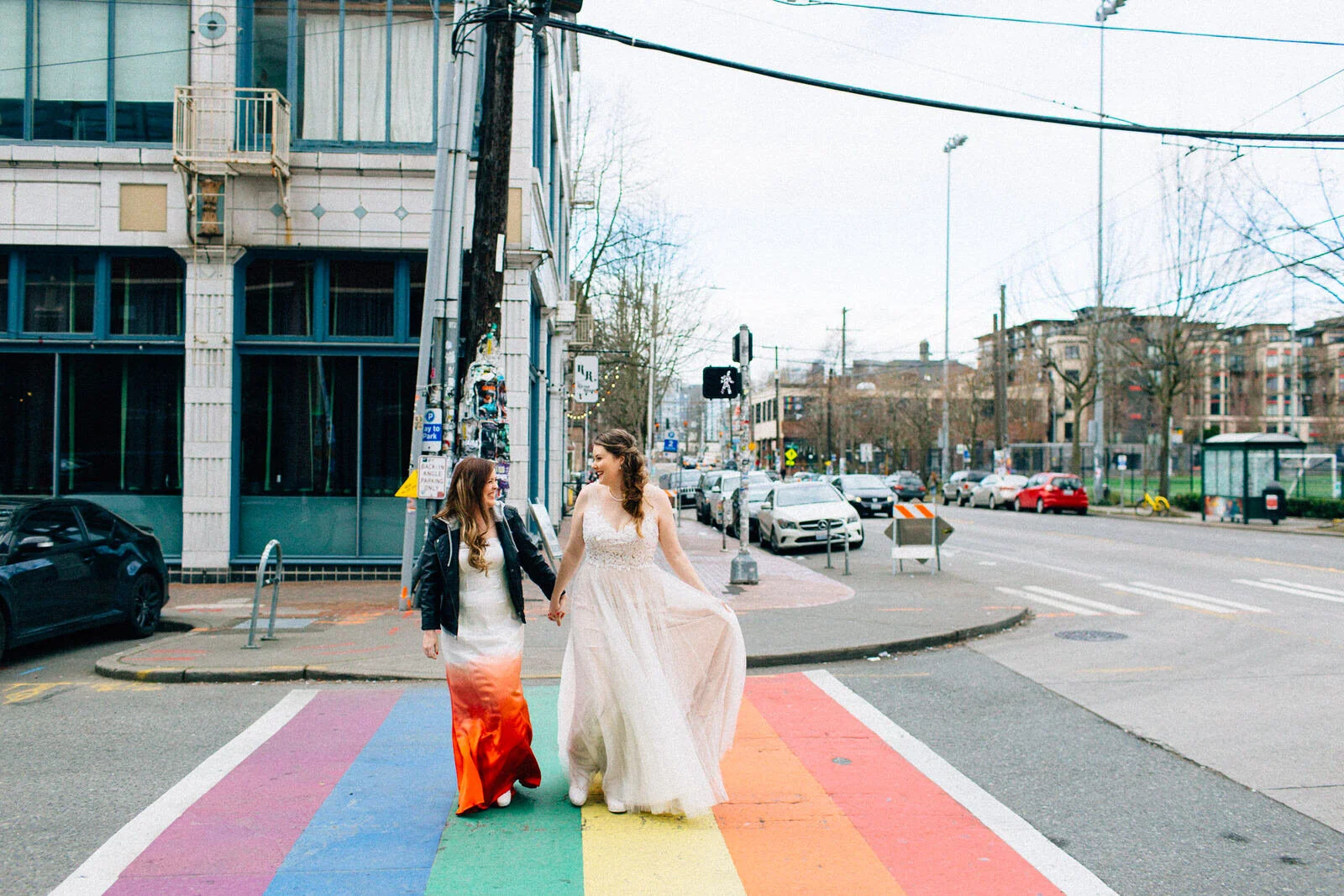 LGBTQ+ couple wearing wedding dresses walks across rainbow crosswalk in Seattle Fuck Yeah Weddings