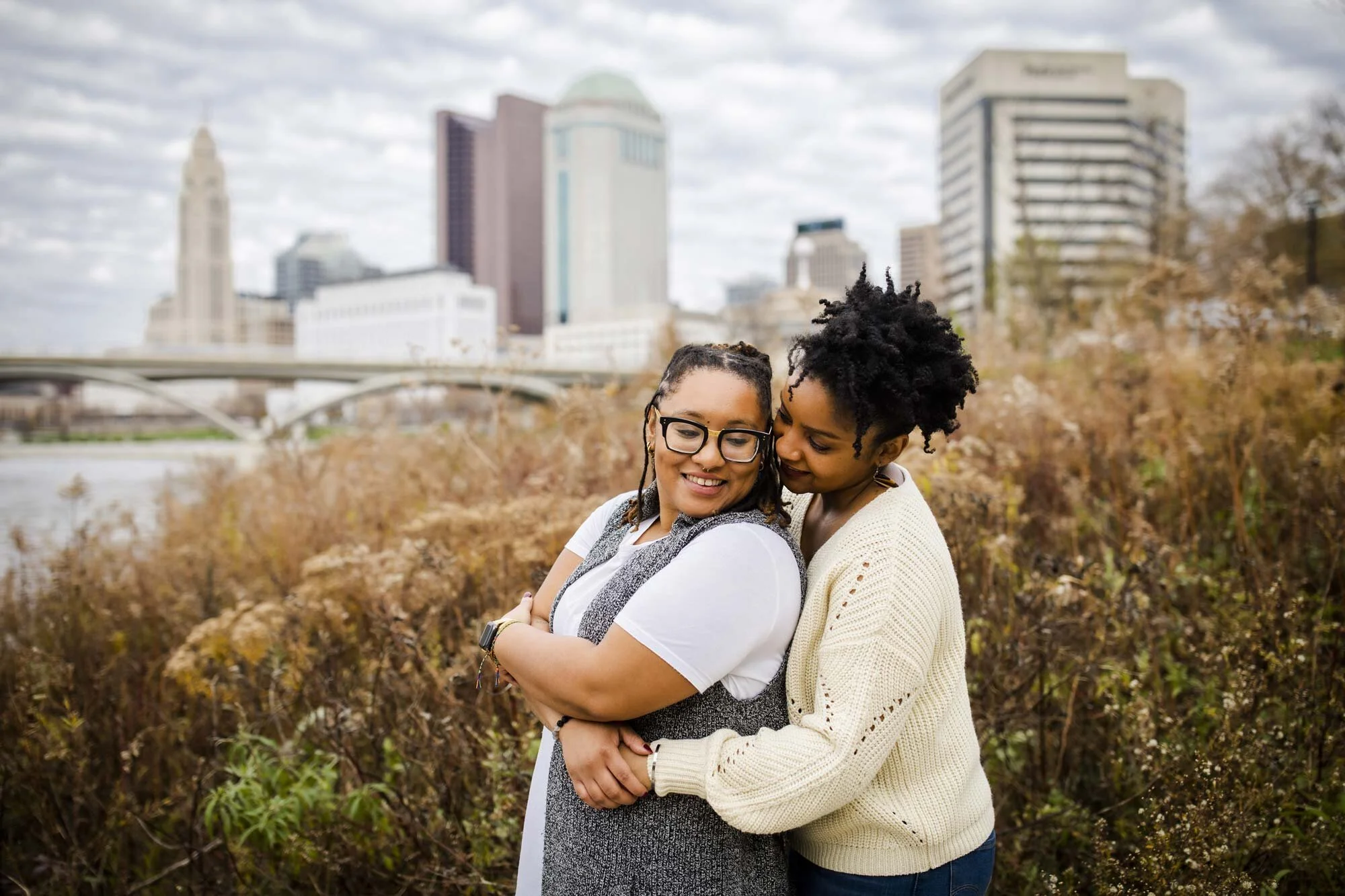 Black couple embraces along waterfront with city skyline behind during LGBTQ Wedding Engagement in Ohio Amy Ann Photography