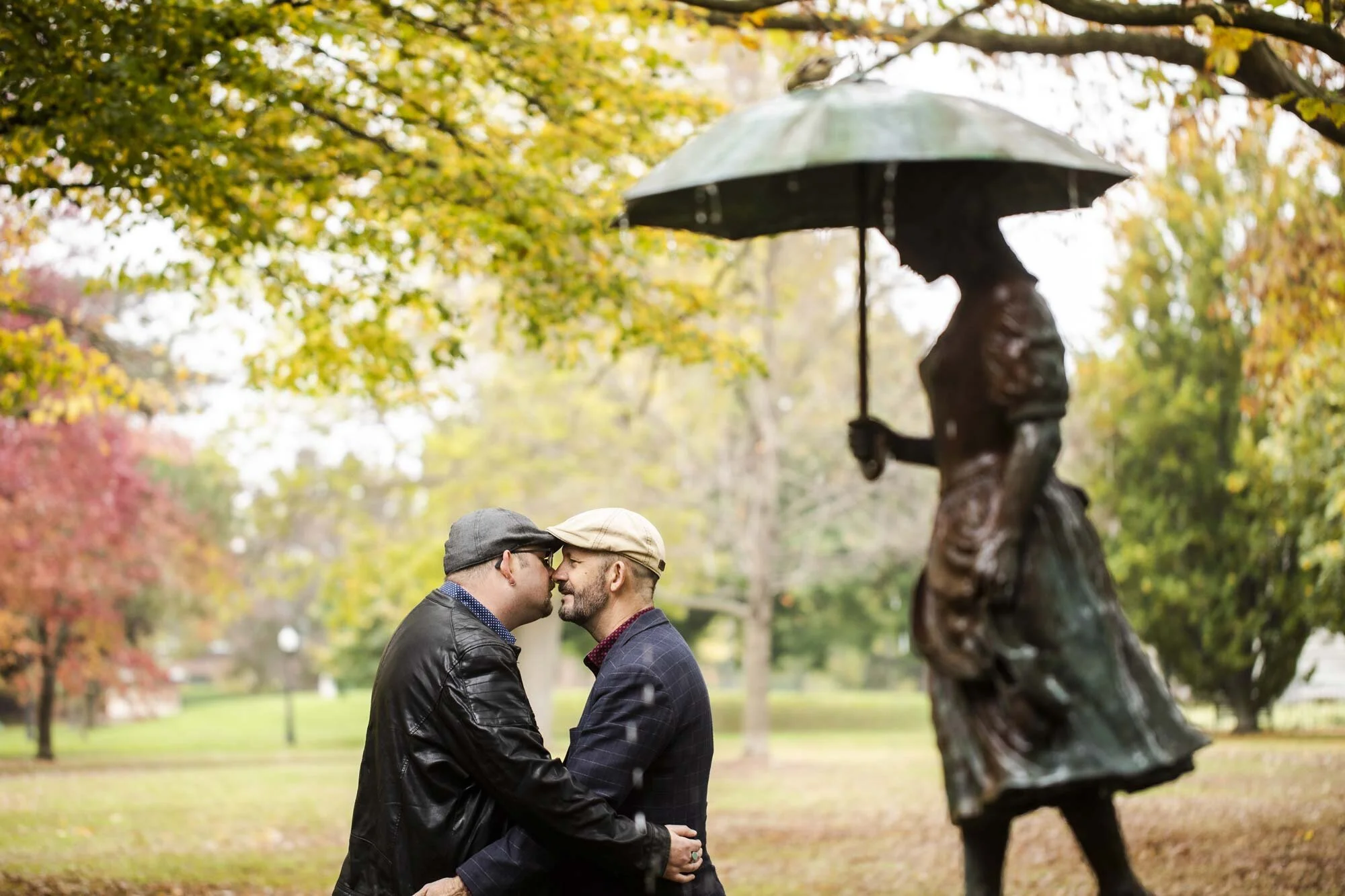 LGBTQ couple in matching hats lean in for a kiss in front of statue during wedding engagement session in Ohio park Amy Ann Photograhy