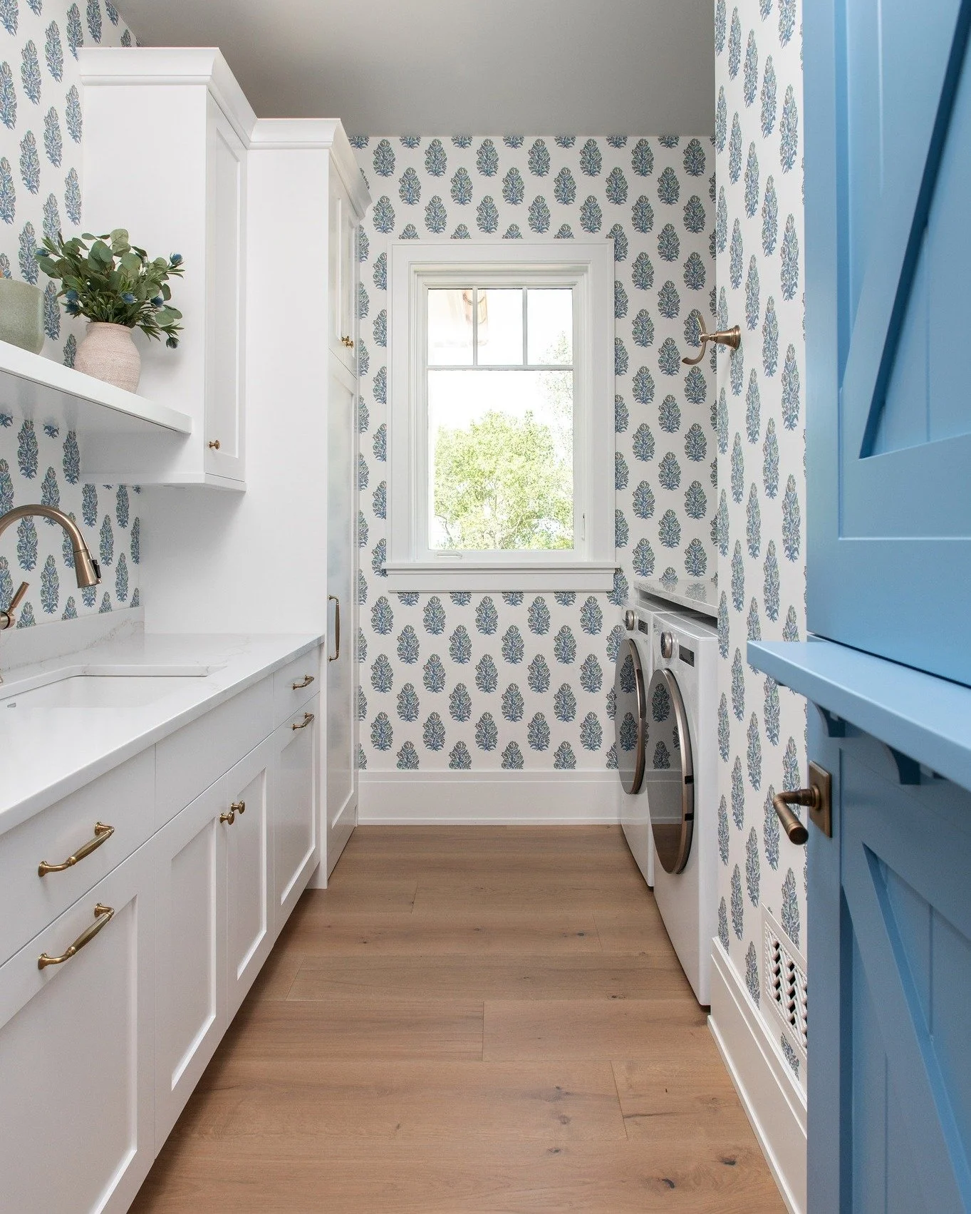 We love giving hardworking spaces just as much personality as the rest of the home. A playful patterned wallpaper brings charm and color to this laundry room, while crisp white cabinetry, warm wood floors, brass details, and that beautiful blue Dutch