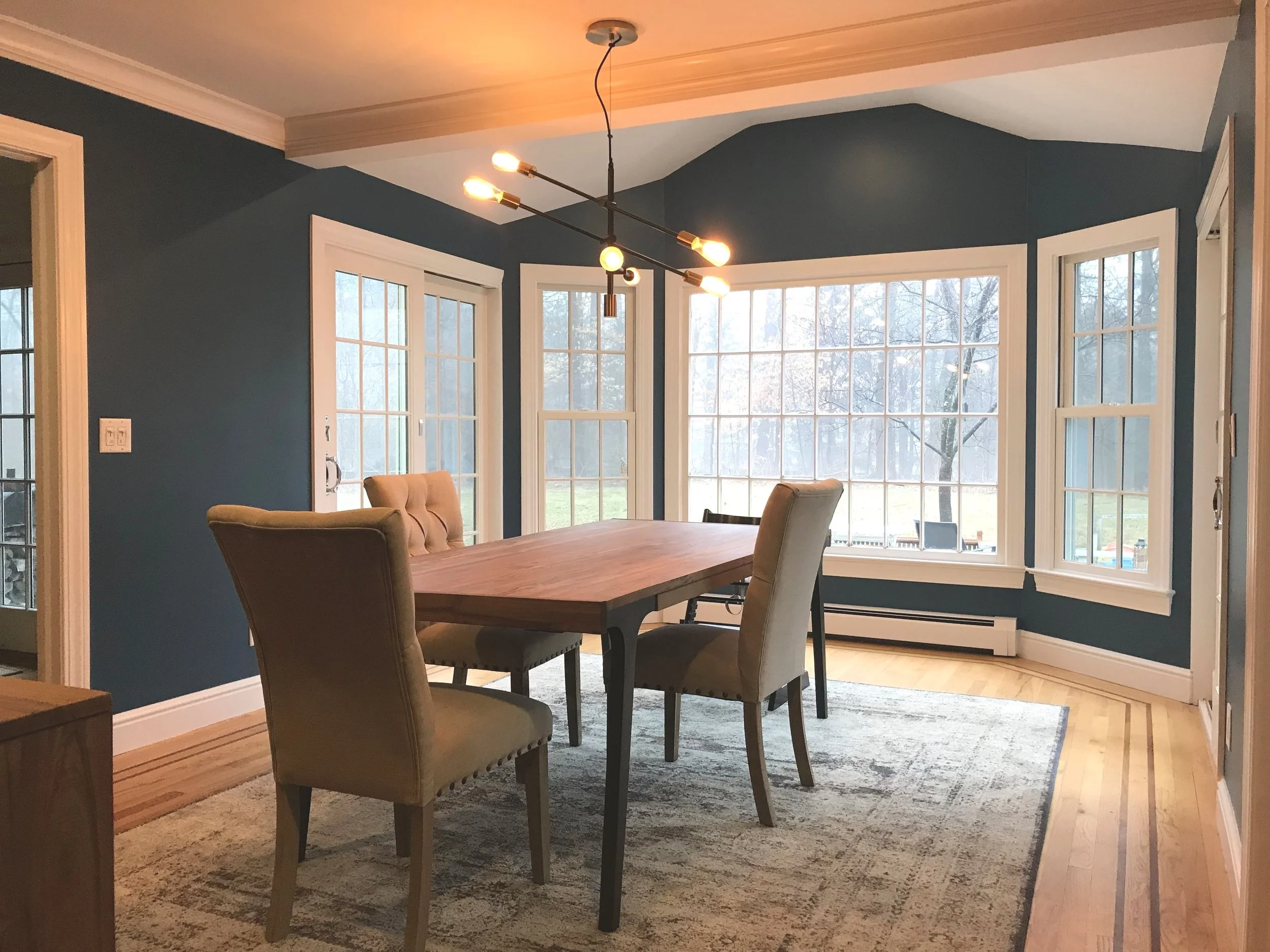 A dining area with a wooden table surrounded by six upholstered chairs. Large windows provide a view of a outdoor area with trees. There is a modern chandelier hanging above the table, and the walls are painted blue with white trim.