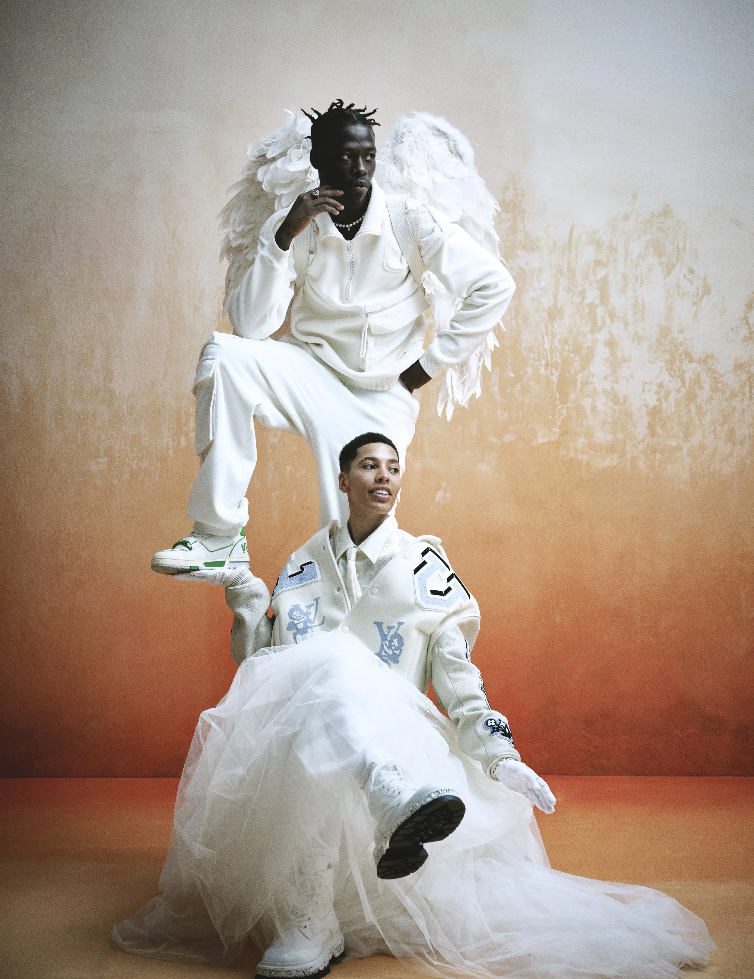Three people dressed in all white with angel wings, posing against a textured peach-colored background. The person on top has dreadlocks, is wearing a white jacket, baggy pants, and sneakers, with angel wings on their back, resting one foot on the sh