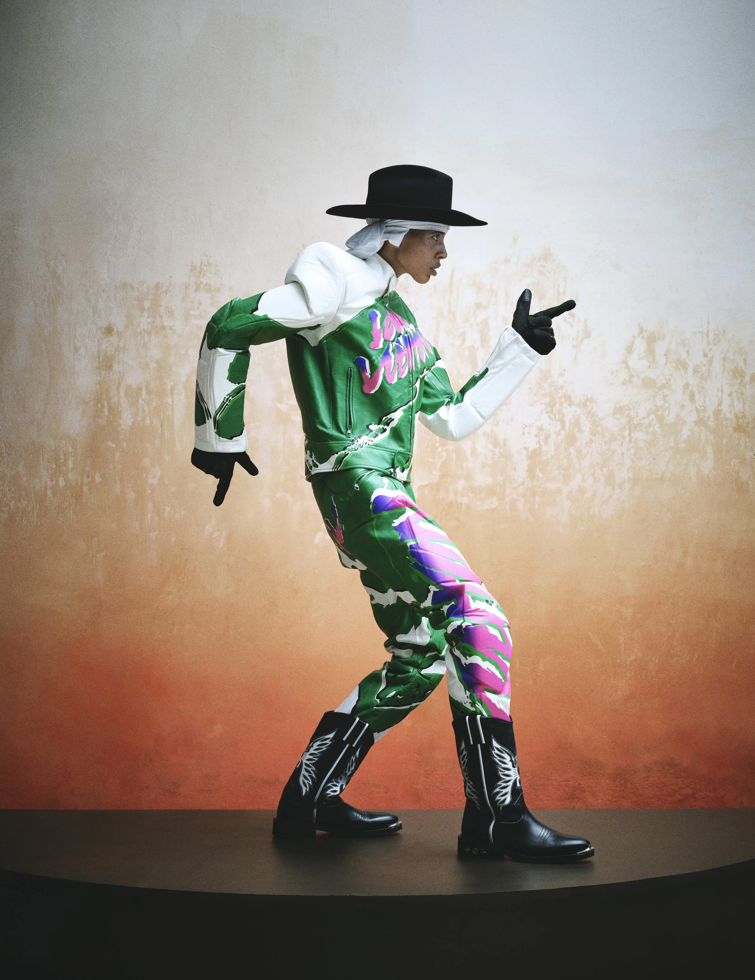 Person wearing a black hat, white shirt, green and pink patterned jacket and pants, standing on stage against an orange gradient background, striking a pose with one hand pointing forward.