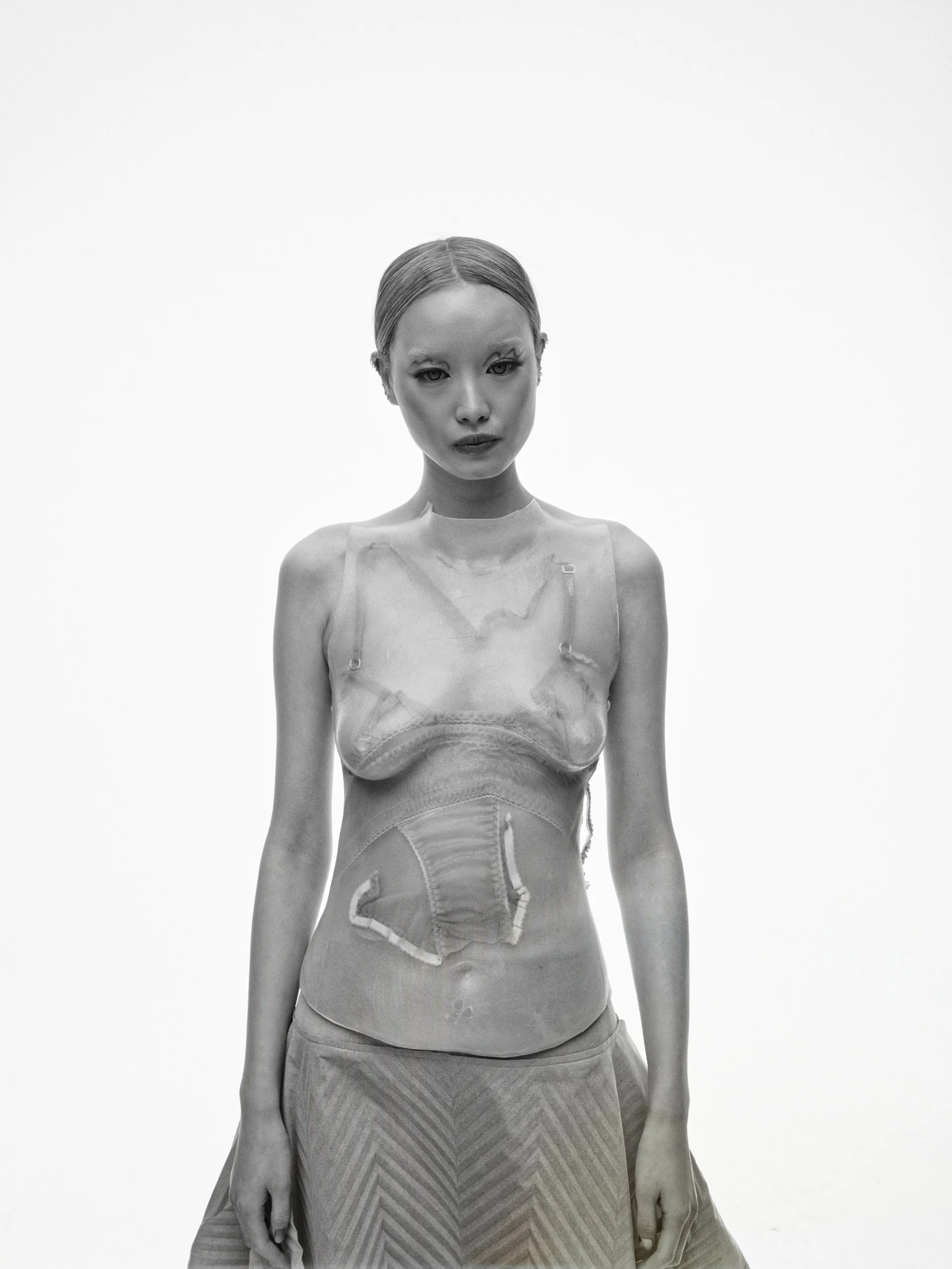 A black and white photo of a woman with short hair, wearing a transparent top and a patterned skirt, standing against a plain background.