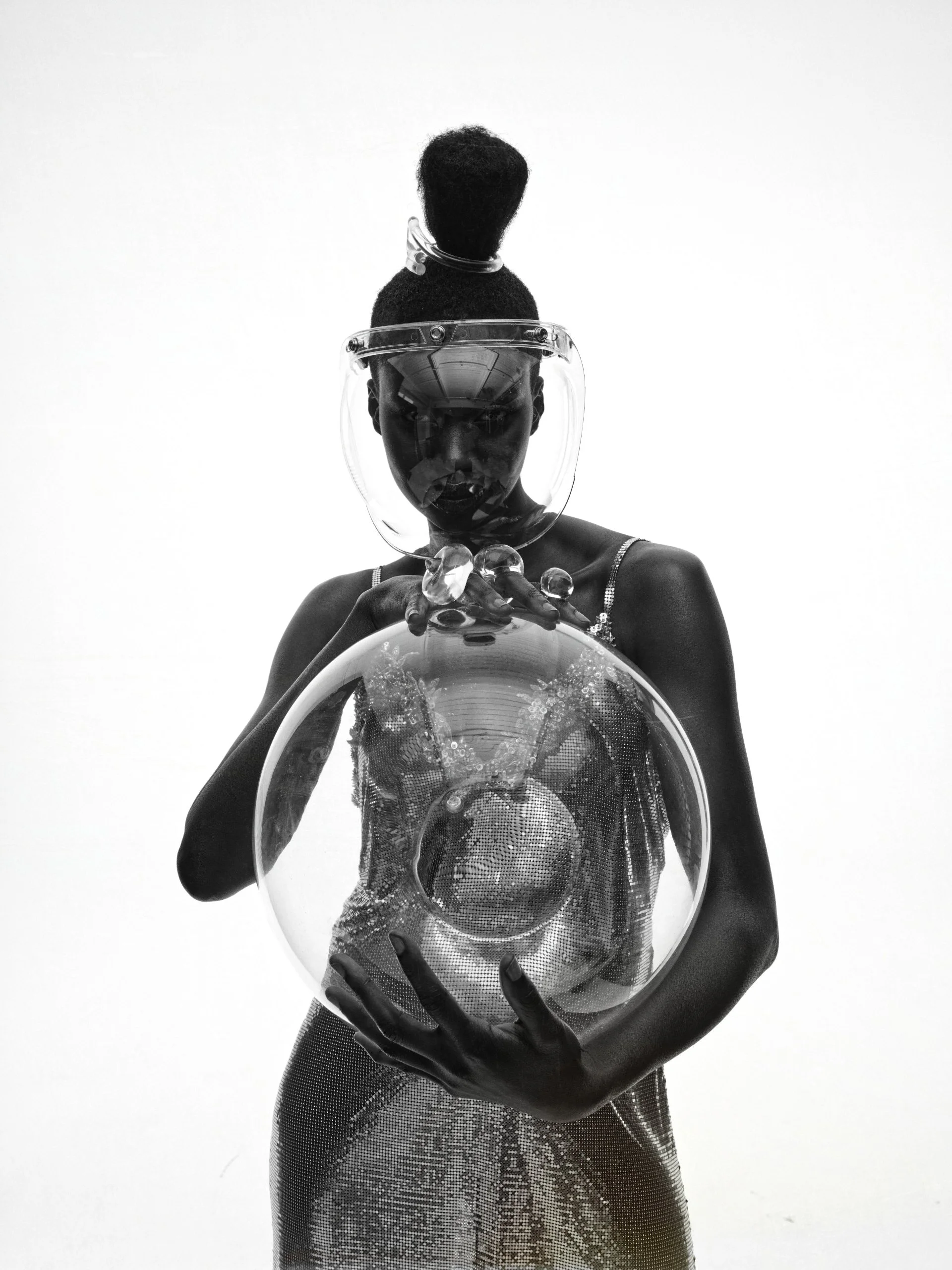 A black and white photo of a person wearing a shiny, patterned dress and holding a large glass bowl with water and bubbles, also wearing a face shield, with their head tilted back and hair styled in an updo.