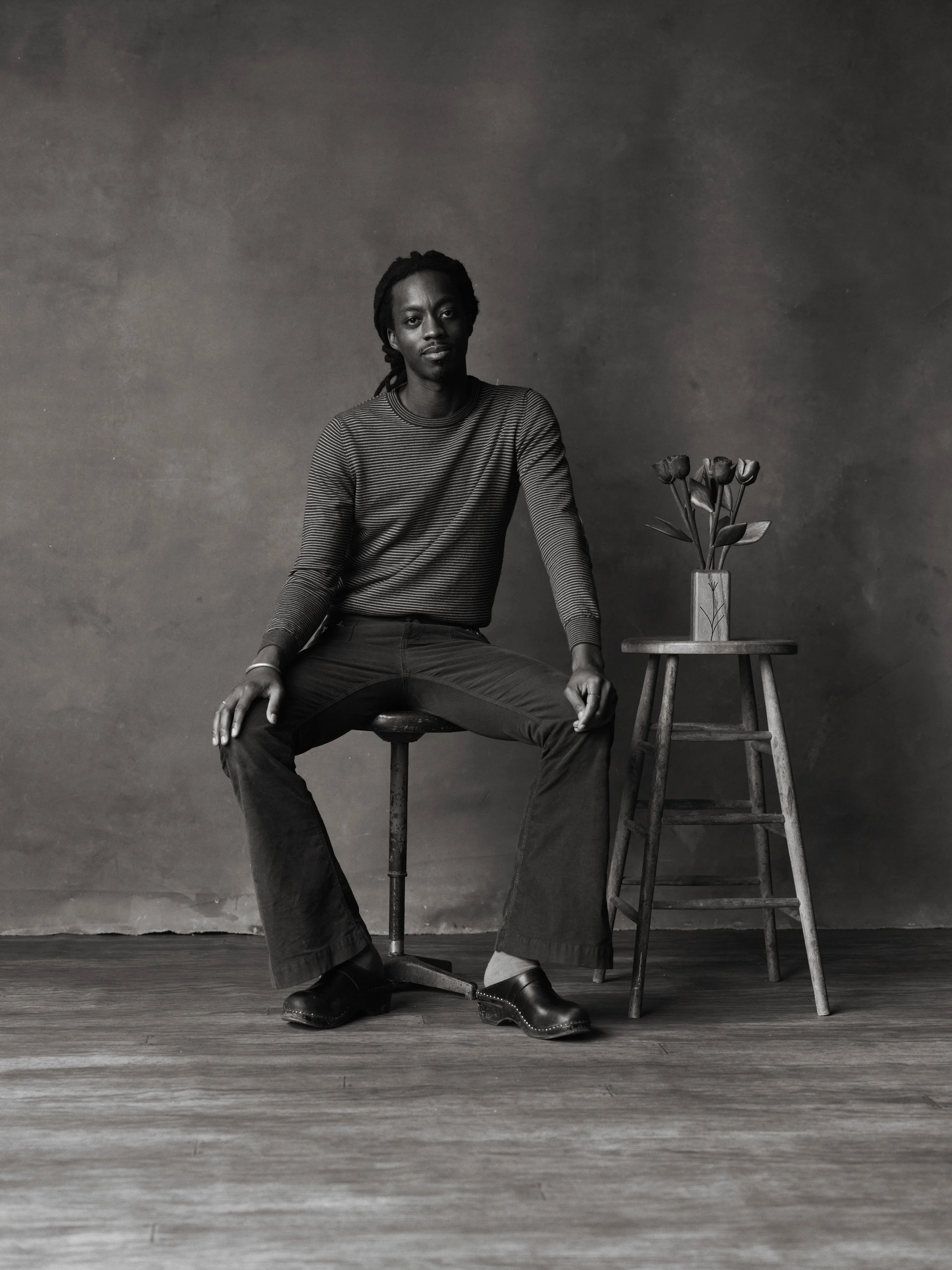 Black and white photo of Justin French with dreadlocks sitting on a stool, next to a small table with a vase of flowers, against a plain wall.