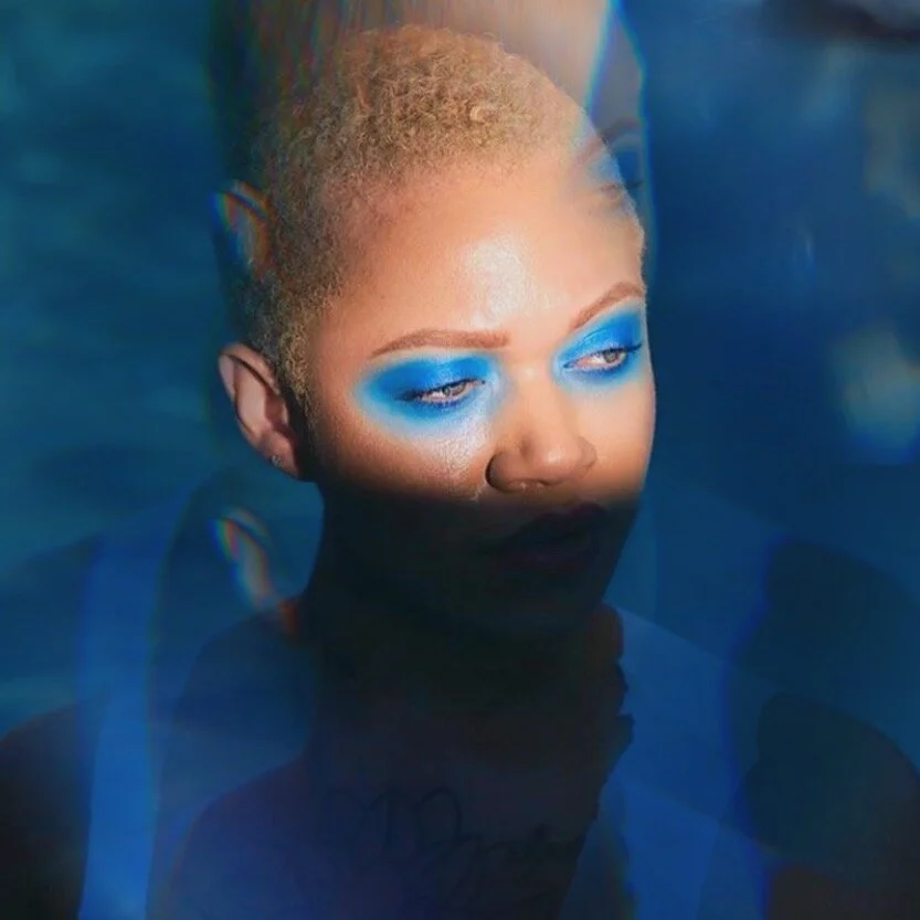A woman with short curly hair and bold blue eyeshadow, with her eyes half-closed, partially submerged in water with a shadow cast across her face.