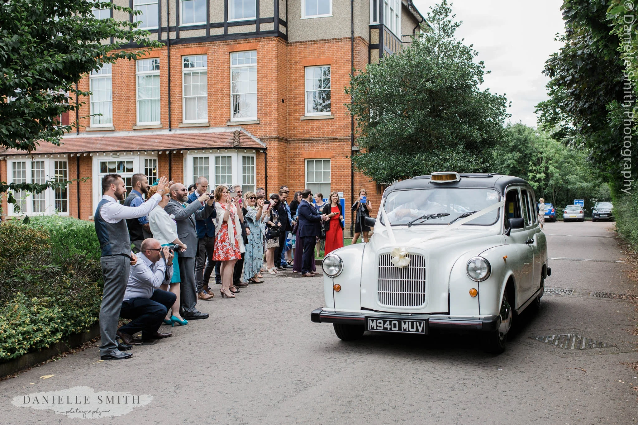 wedding guests waving off bride and groom in white taxi