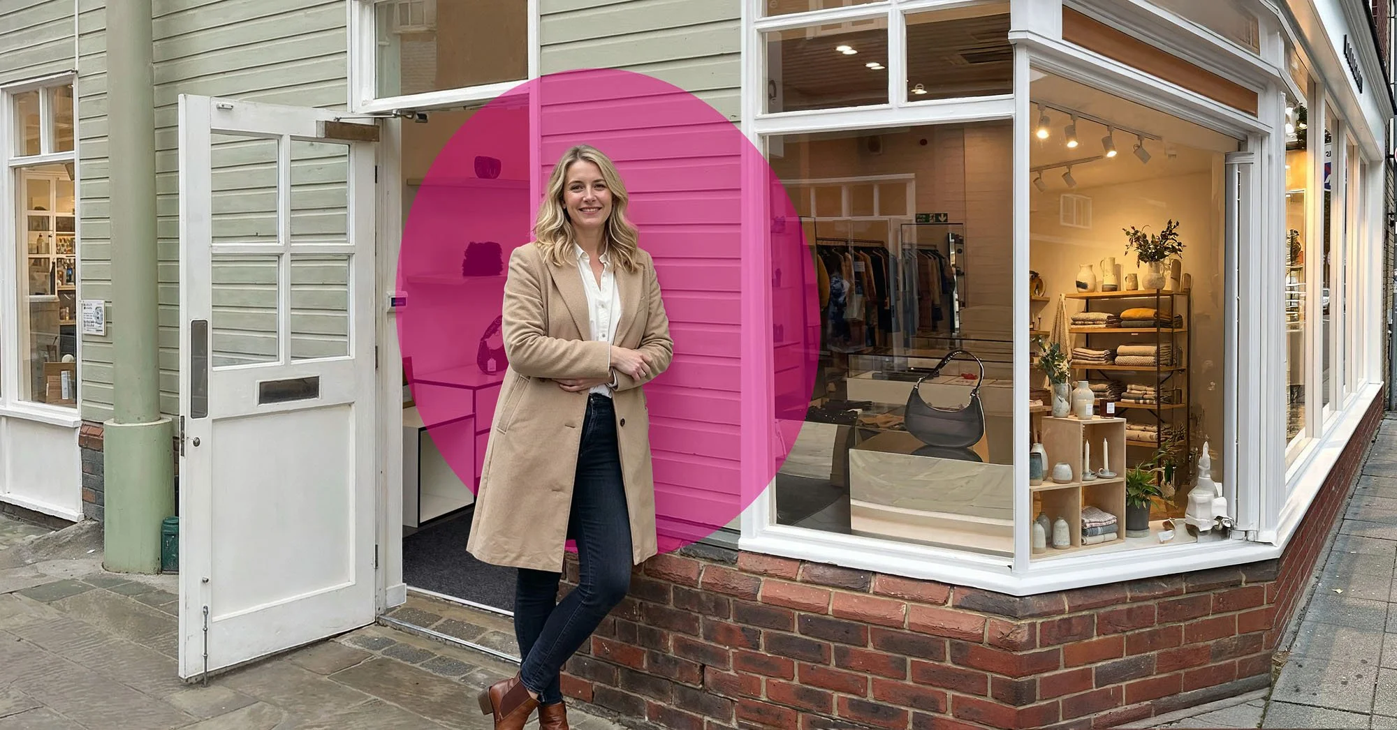 Woman standing outside a boutique store with large glass windows, beige coat, black jeans, and brown boots, smiling and posing in front of the store.