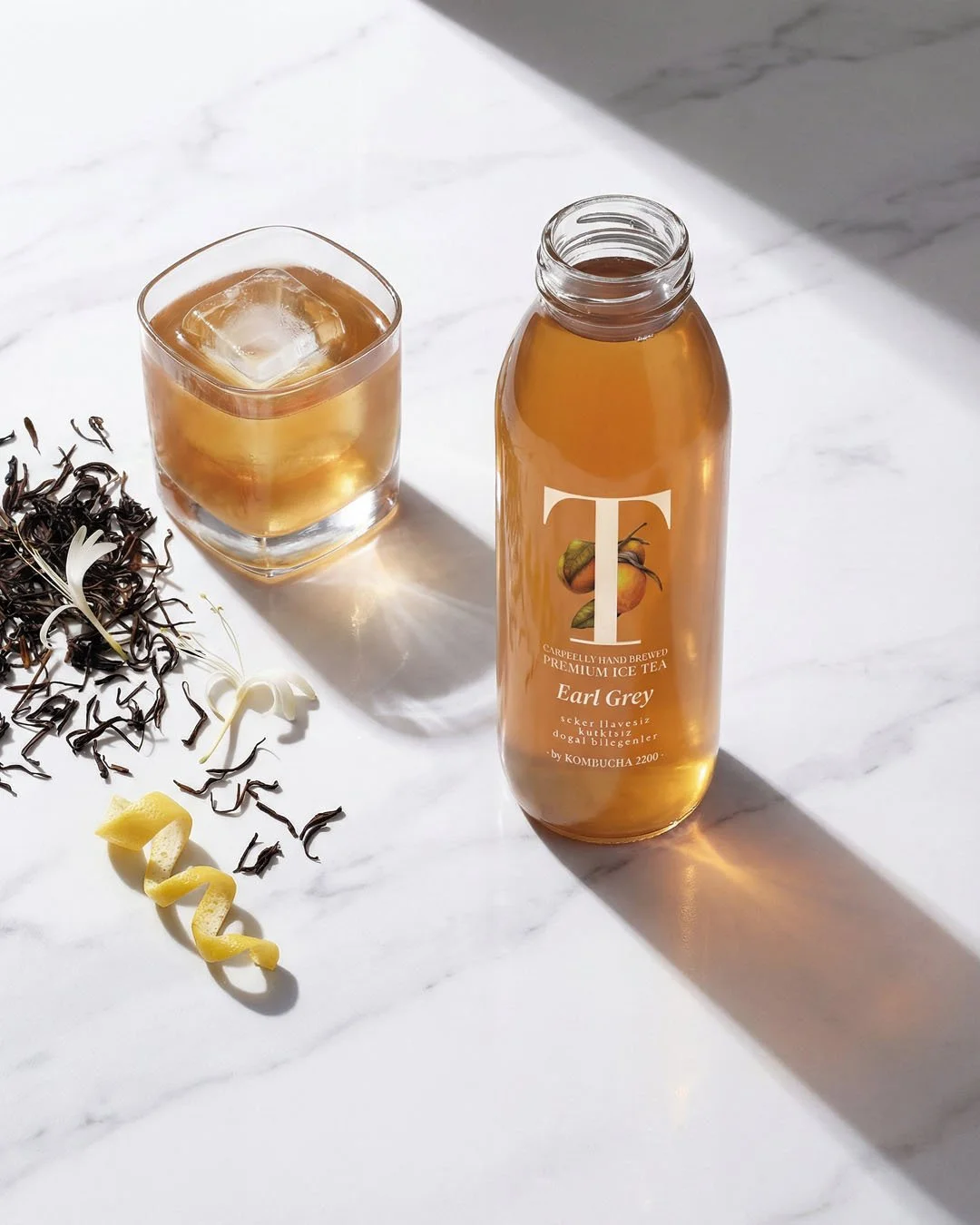 A bottle of Earl Grey flavored iced tea next to a glass with ice and tea, on a white marble surface with loose tea leaves and lemon peel scattered nearby.