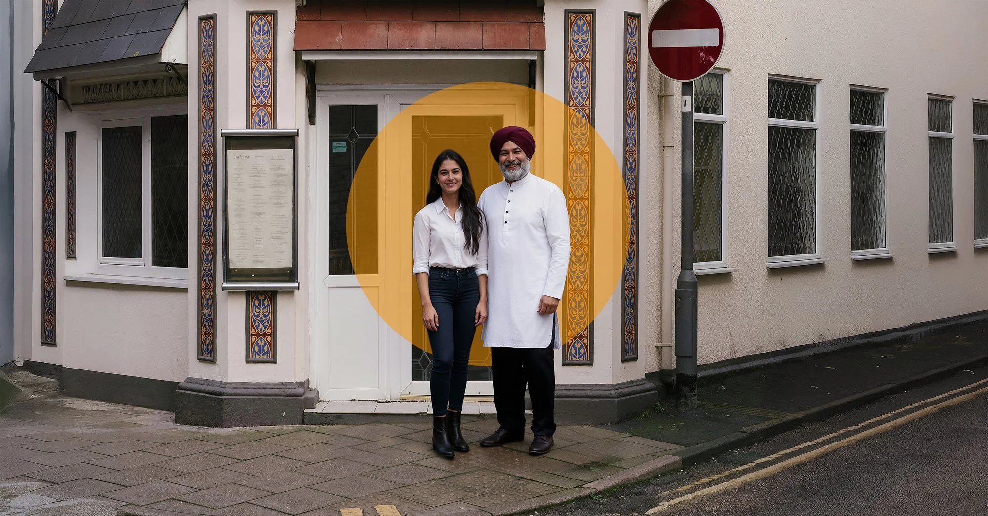 A woman and a man standing outside a building with a decorative tile border. The man is wearing a white traditional outfit and a maroon turban, while the woman is dressed in a white blouse and dark pants.