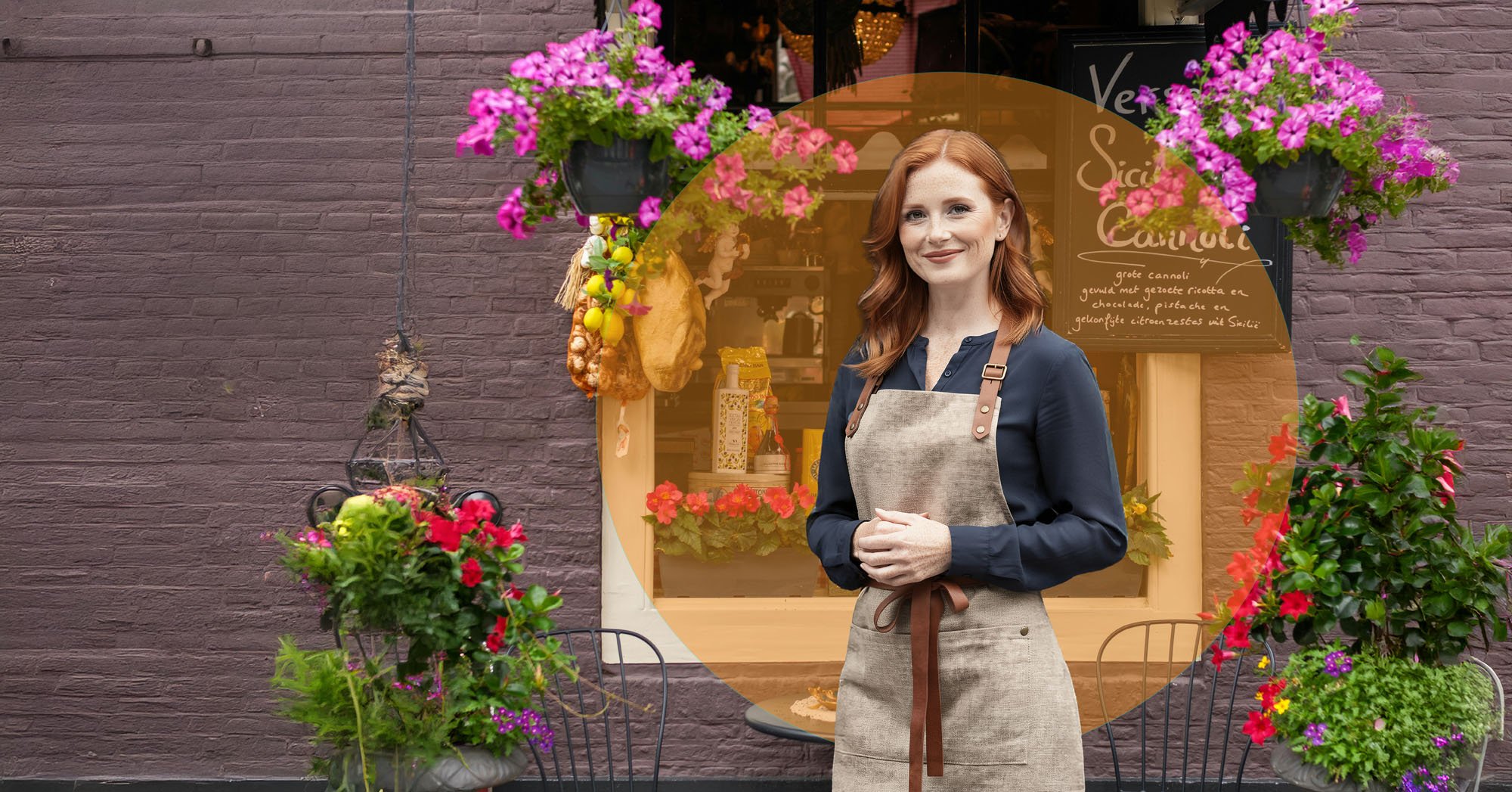 A woman standing outside a flower shop, wearing an apron, smiling, with pink and purple flowers hanging above and placed in planters around her.