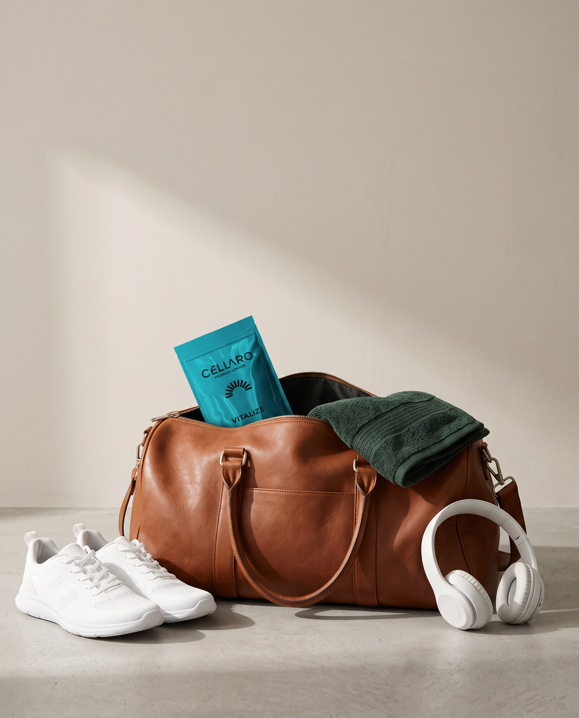 Travel or workout essentials including white athletic shoes, a tan leather duffle bag, a green towel, a blue protein shake packet labeled 'CELLAR VITALIZE,' white headphones, and a white suitcase or object partially visible on a light grey floor agai