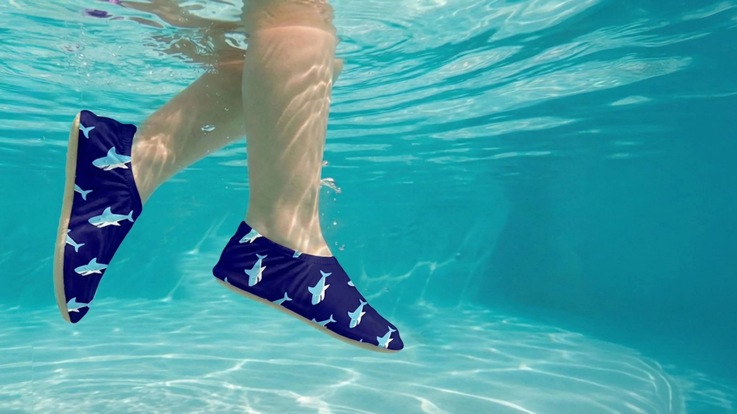Child wearing blue swim trunks with shark pattern, underwater in a pool.