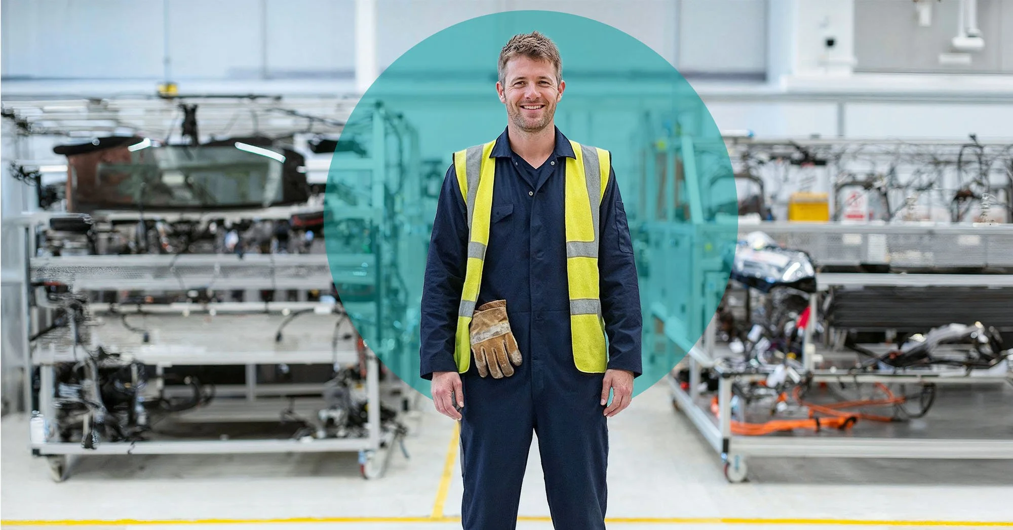 A male worker in safety gear, including a yellow safety vest and brown gloves, standing in an industrial or manufacturing setting with metal equipment and machinery in the background.