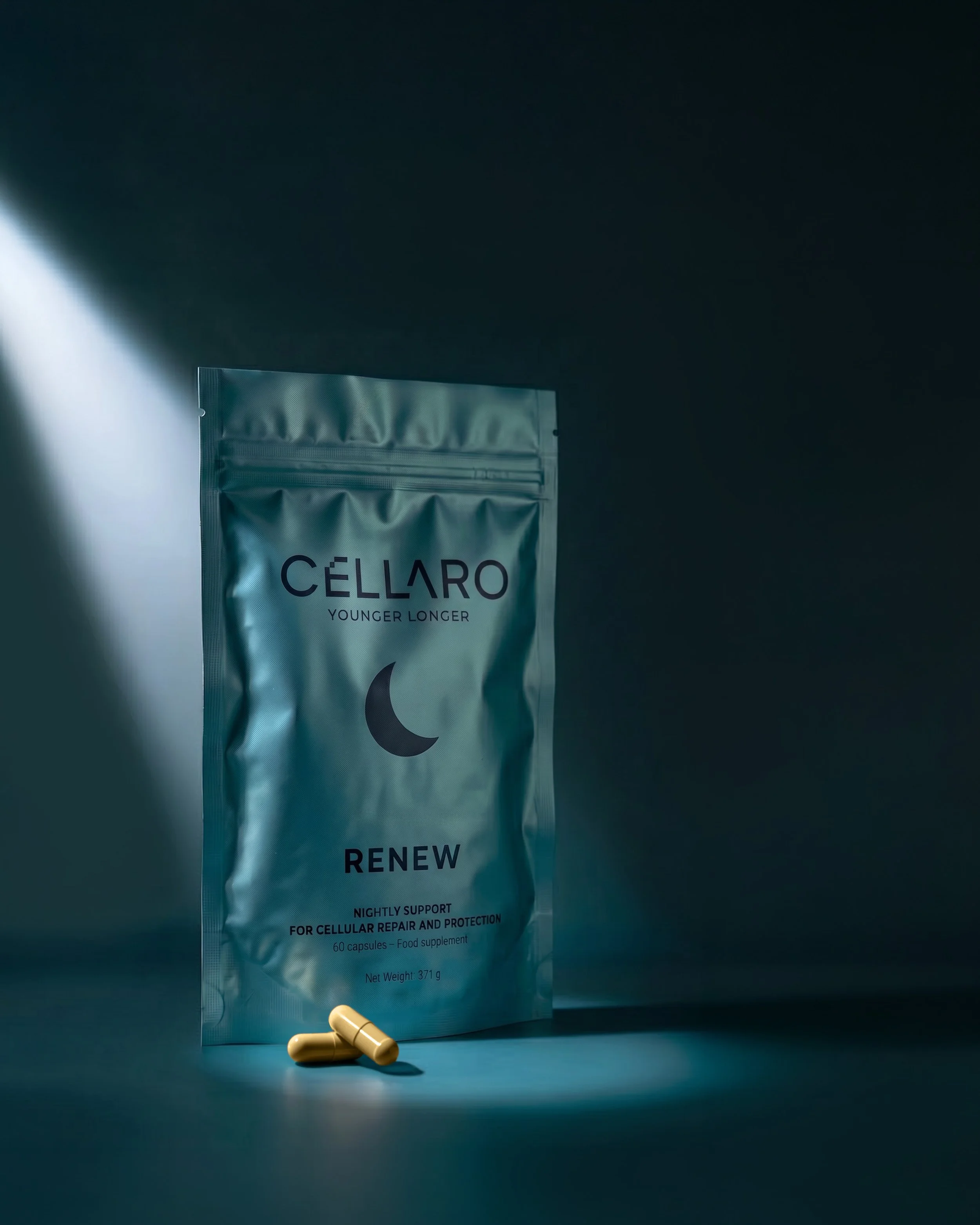 A metallic package labeled 'CELLARO' with the slogan 'YOUNGER LONGER,' standing upright on a dark surface, with two beige capsules in front of it, and additional small capsules near the bottom of the package.