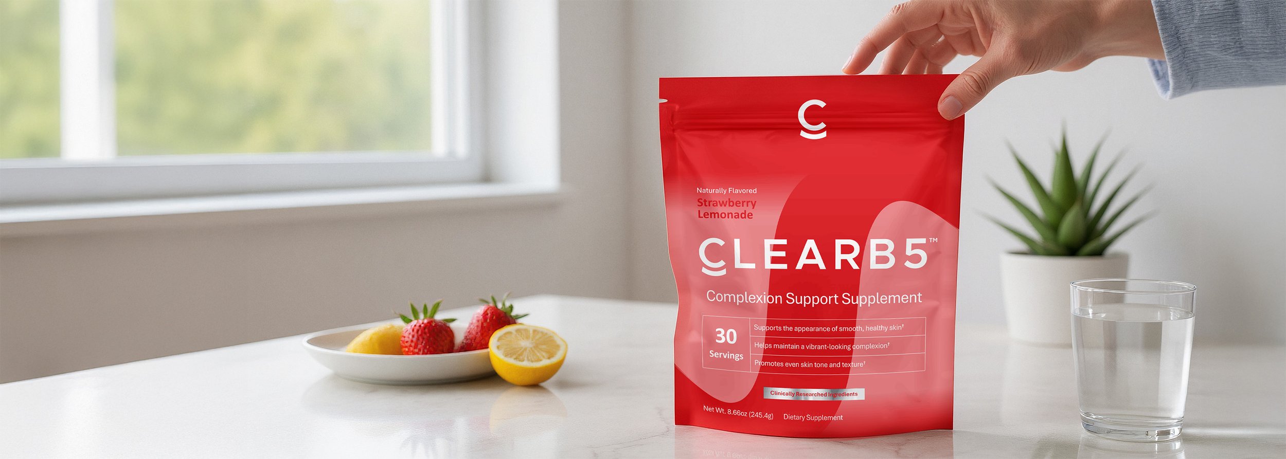 A person holding a red pouch of Clearb5 supplement on a white countertop with a bowl of strawberries and lemon slices, and a glass of water nearby, in a bright room with a window and a small potted plant in the background.