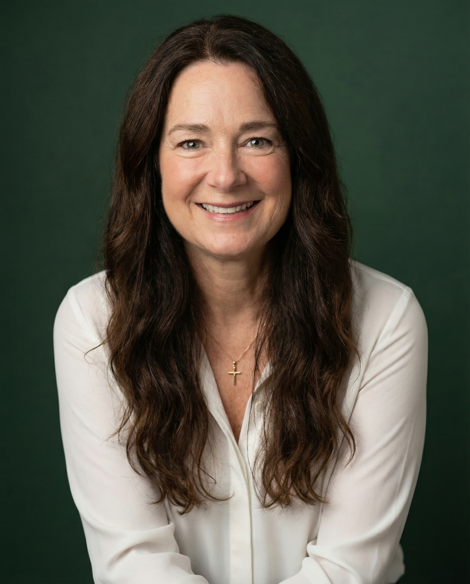 A woman with long brown hair wearing a white blouse and a cross necklace, smiling with a green background.