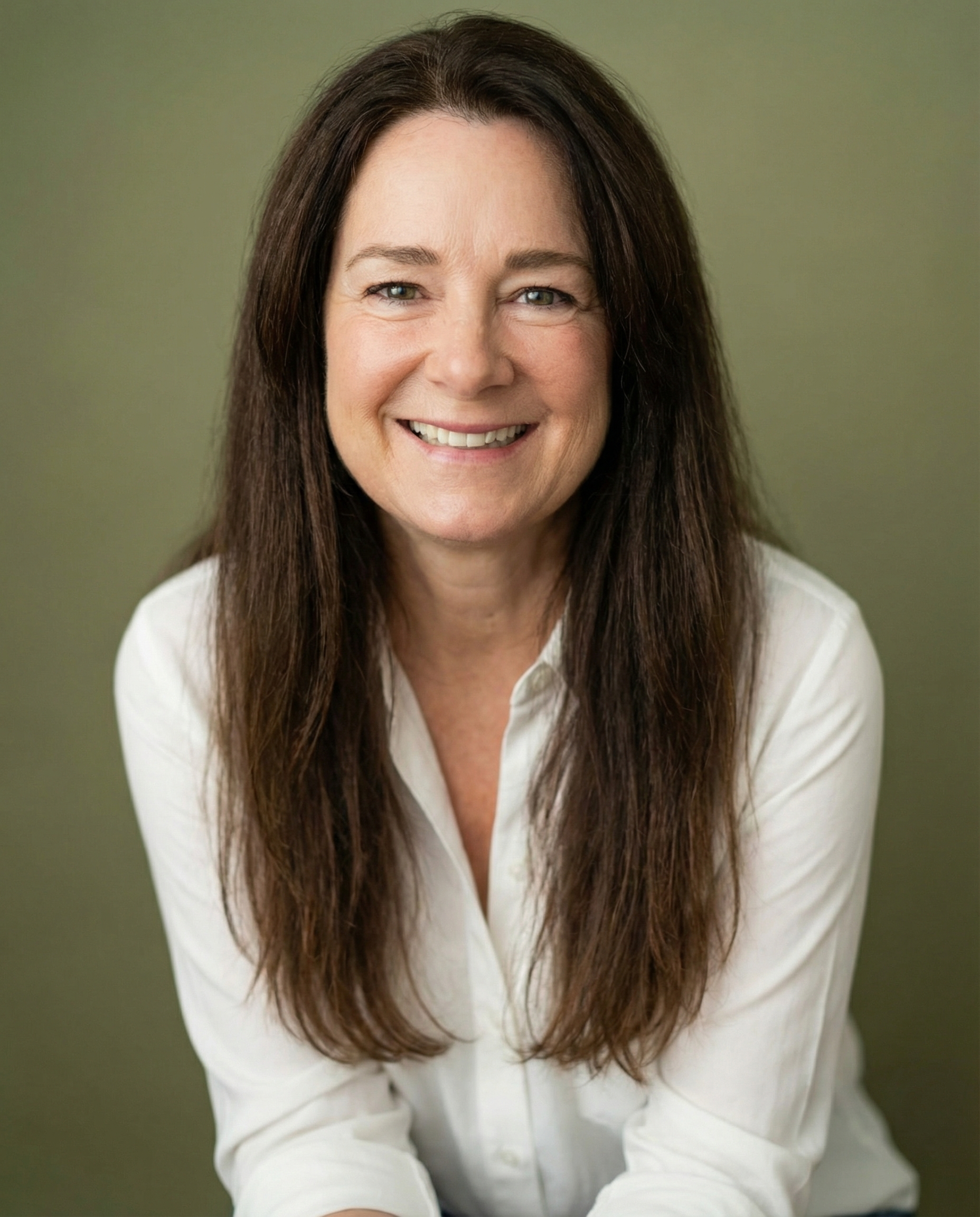 A middle-aged woman with long brown hair, wearing a white button-up shirt, smiling in front of a plain olive green background.