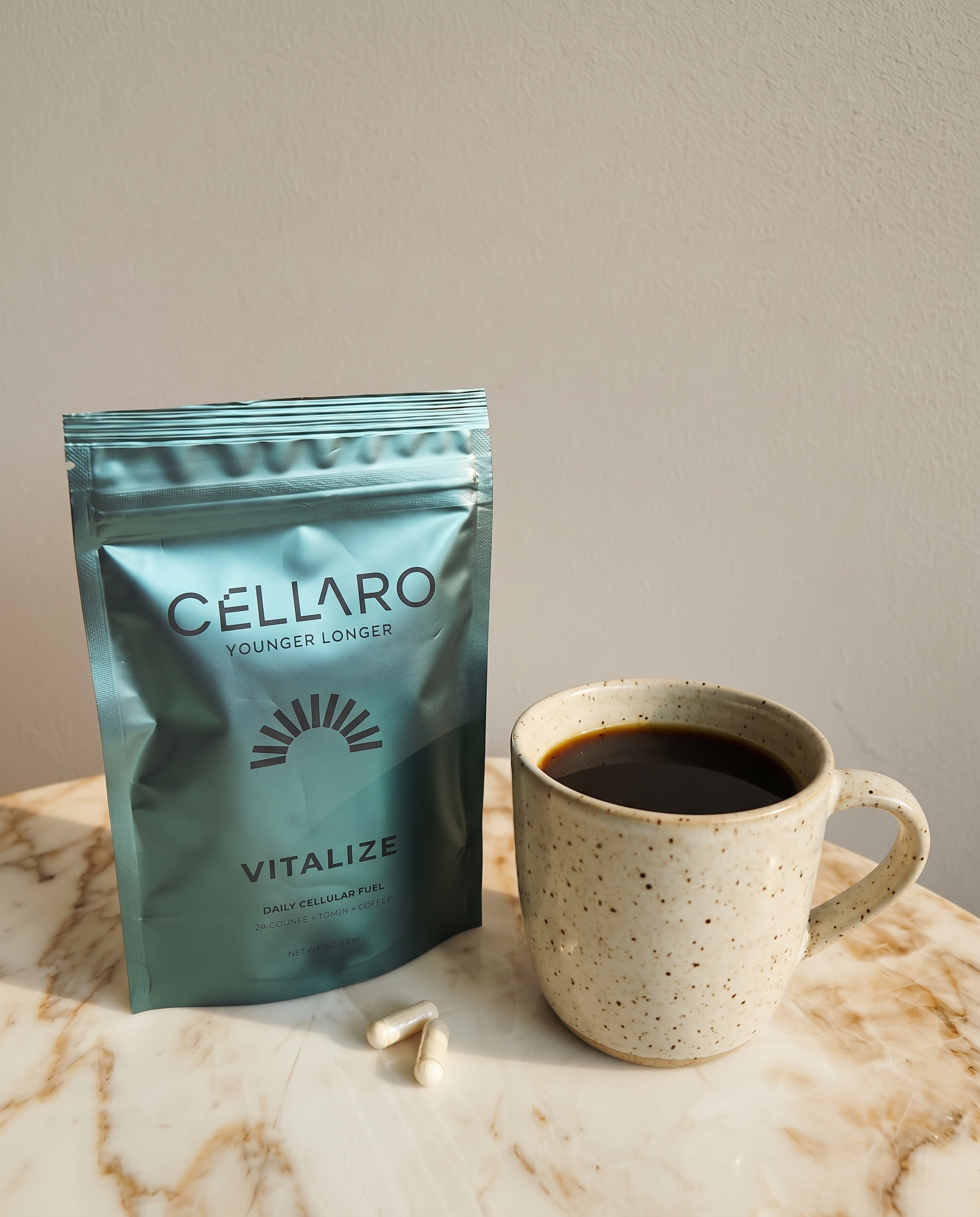 A marble countertop with a speckled ceramic mug filled with black coffee, a blue bag labeled 'Cellaro Vitalize Younger Longer,' a few white capsules, and a white spoon.
