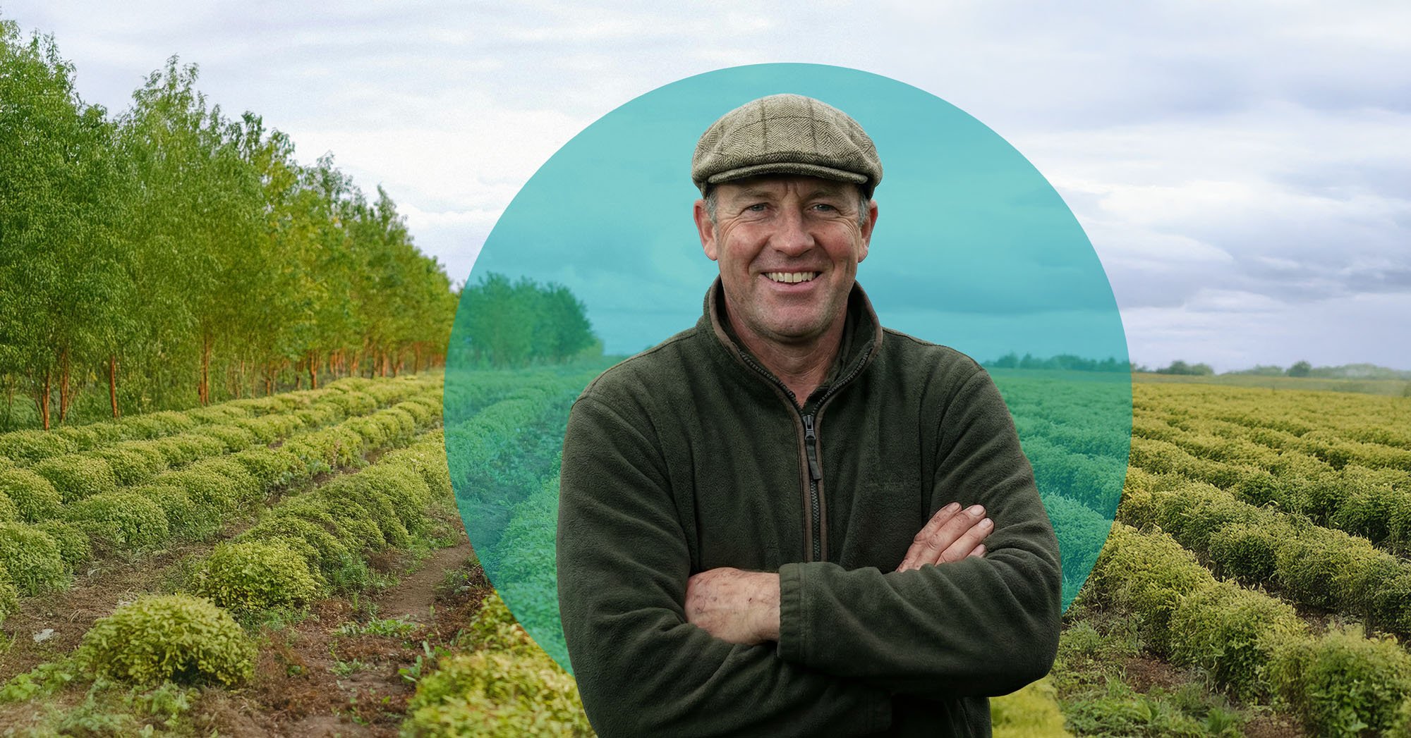 A man in a green jacket and a flat cap smiling with crossed arms standing in a field of cultivated bushes with green trees and a cloudy sky in the background.