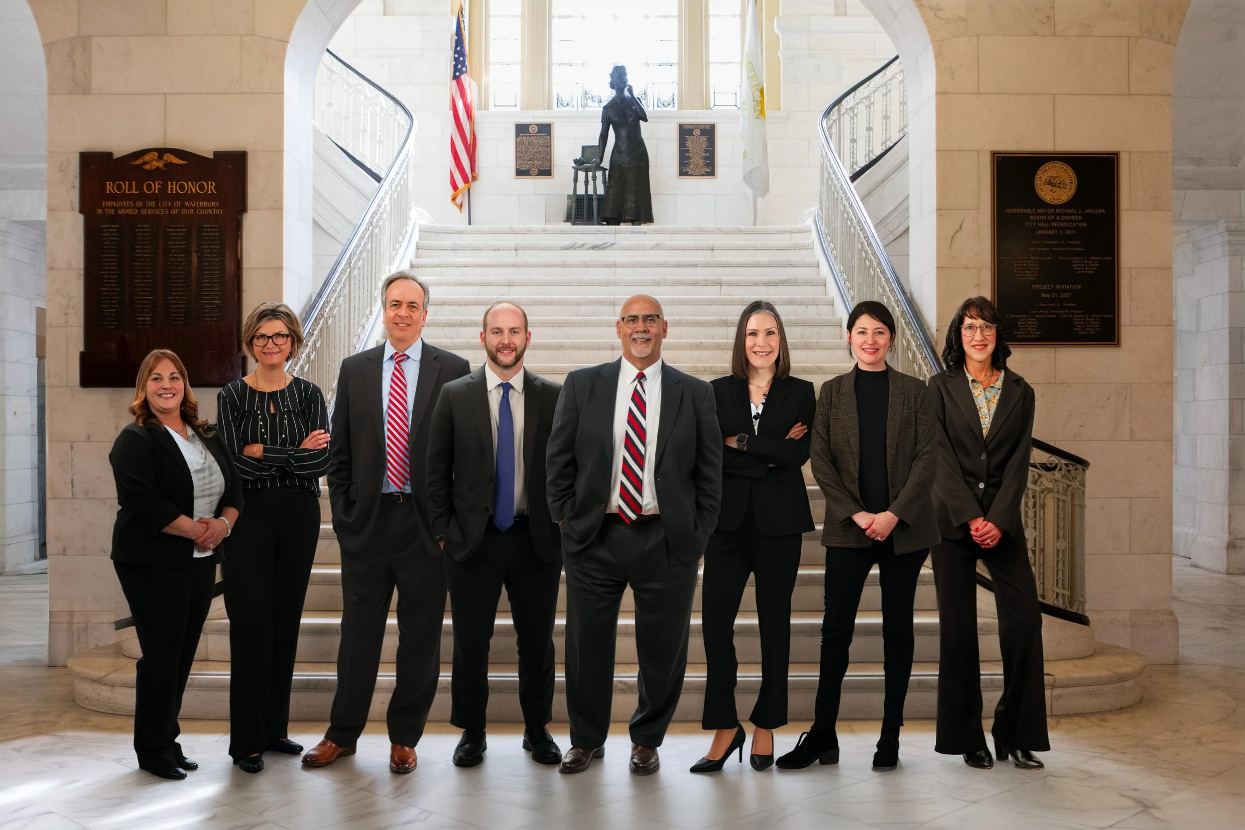 Group of eight professionally dressed individuals standing in front of a staircase inside a government building, with plaques and flags behind them.