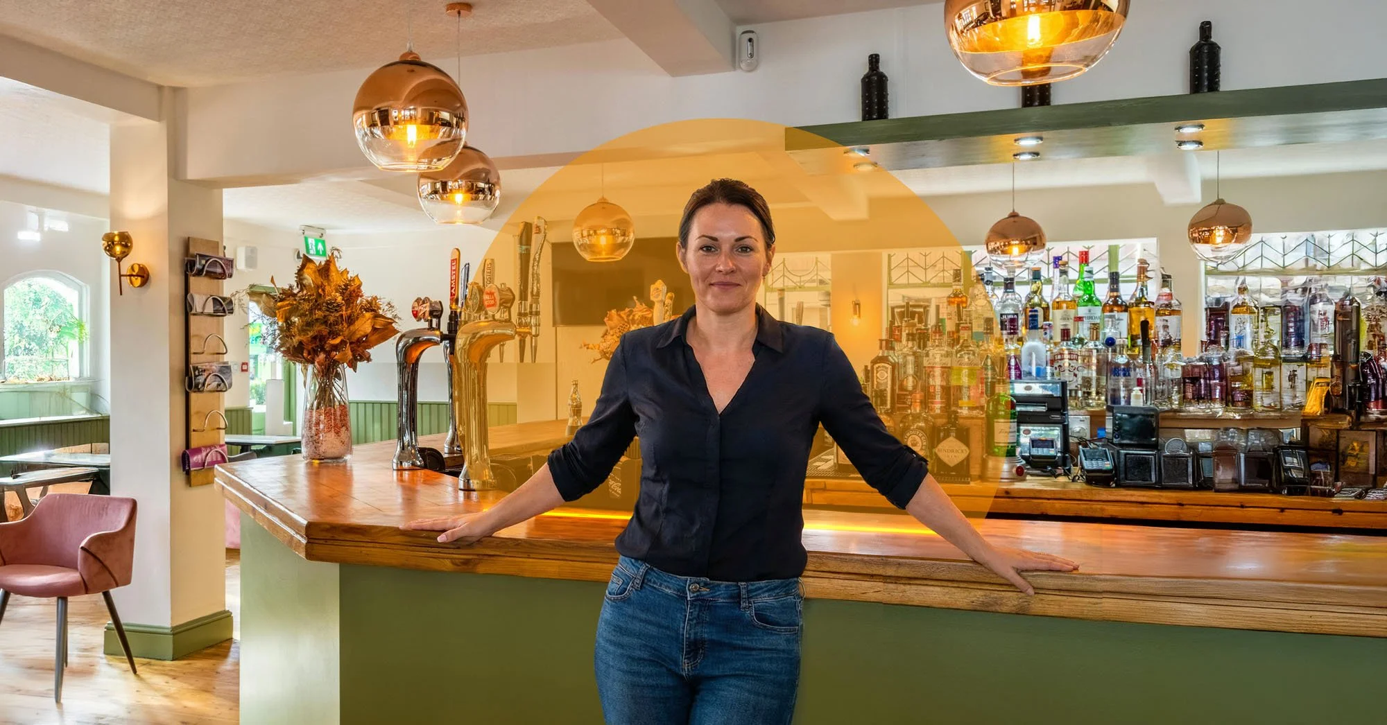 A woman standing behind a bar counter in a restaurant or bar, with bottles and taps visible behind her, and warm lighting overhead.