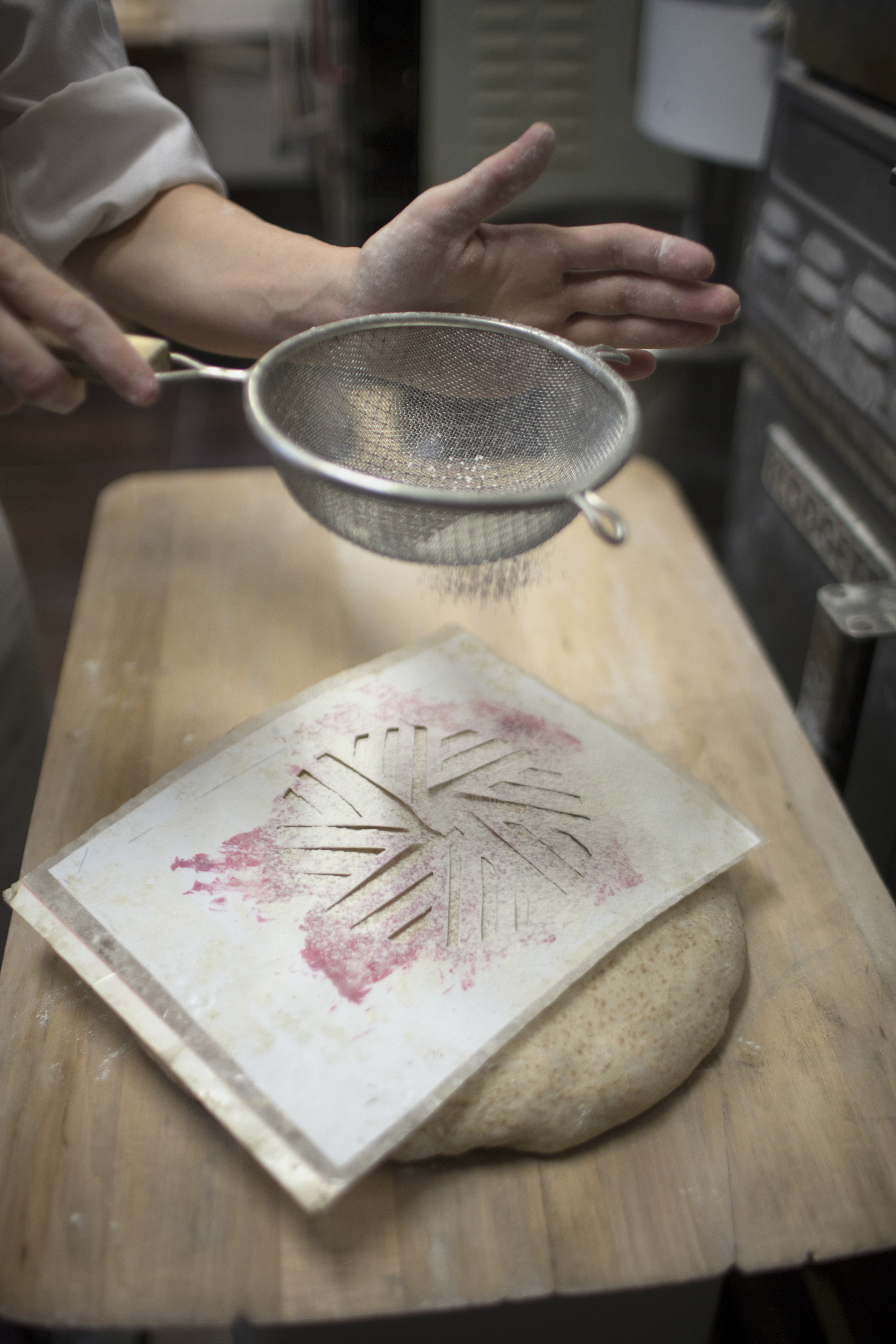 flour being dusted through a template of the runner and stone logo onto the top of the bolzano rye miche in the bakery