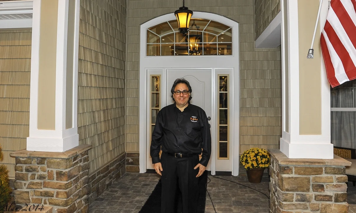 Pat Perrino standing at the entrance of a custom-built home with stone pillars, shake siding, and decorative exterior lighting.