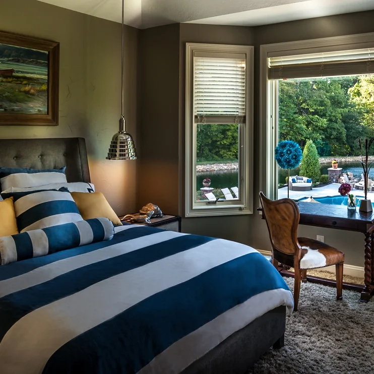Cozy bedroom with large windows, striped bedding, modern pendant light, and scenic view of a backyard pool and greenery.