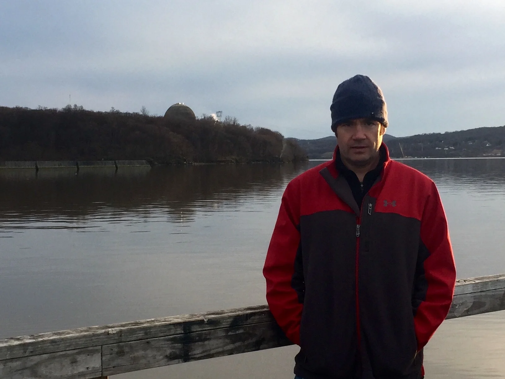 Christopher Swain stands beside the Hudson River in Peekskill, New York in March 2016. The Indian Point Energy Center nuclear power facility is partially visible on the hill behind him. (This image belongs to Christopher Swain. Members of the news media: you are welcome to use this image.)