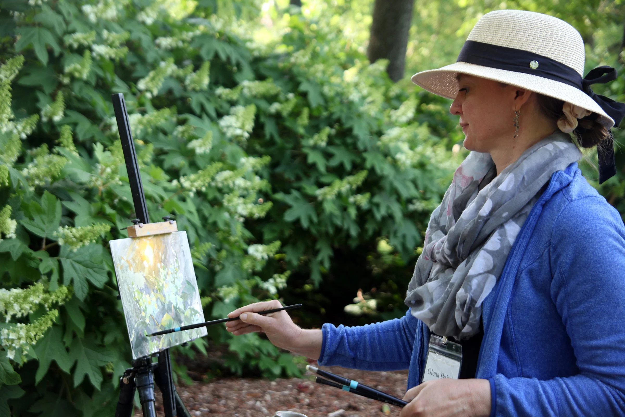 Woman in a wide-brimmed hat and scarf painting en plein air outdoors with a palette and brushes.