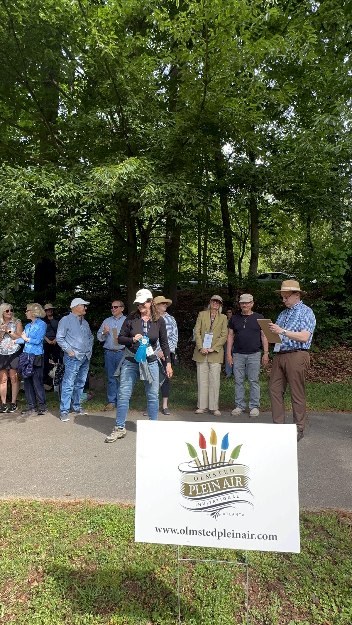 Group of people gathered outdoors near a sign for the Olmsted Plein Air Invitational in Atlanta, with trees in the background.