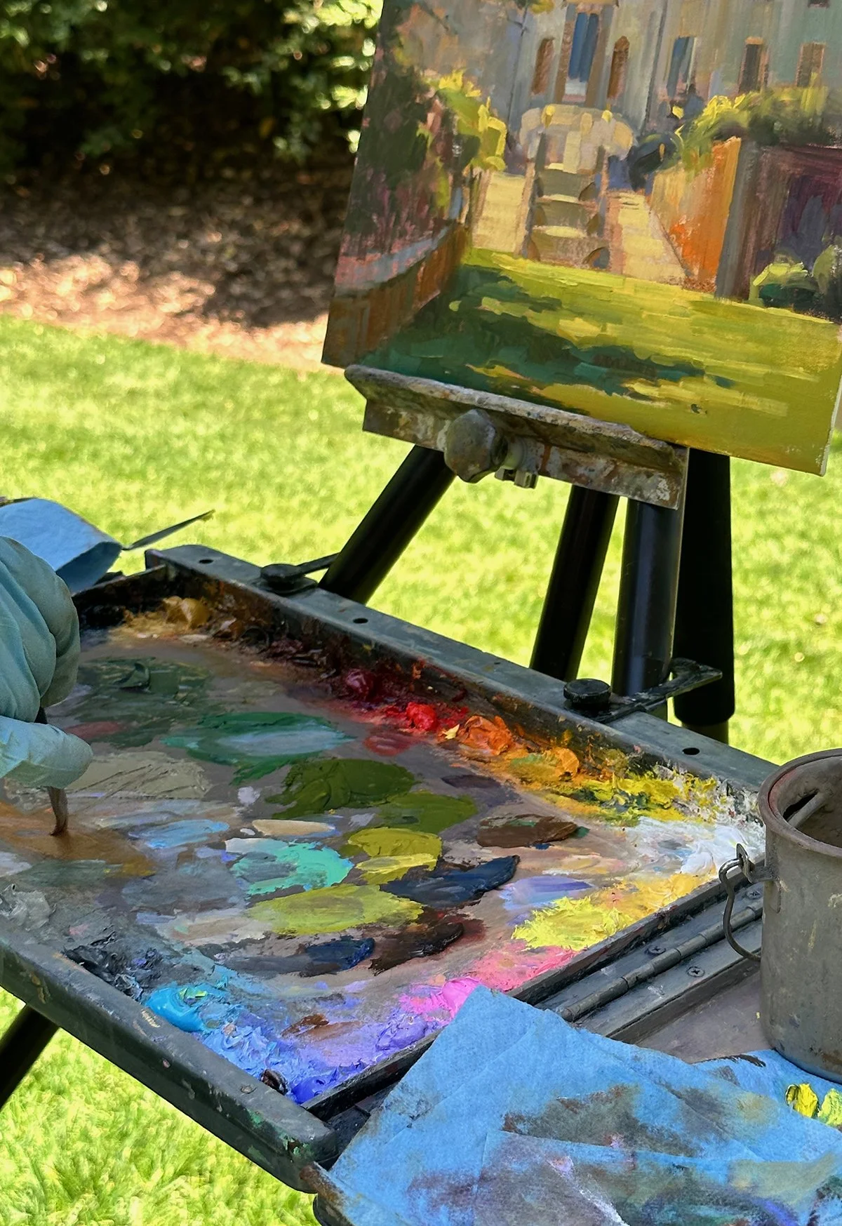 Painter's palette with mixed colors and a small canvas on an outdoor easel depicting a colorful street scene with buildings and trees in the background.