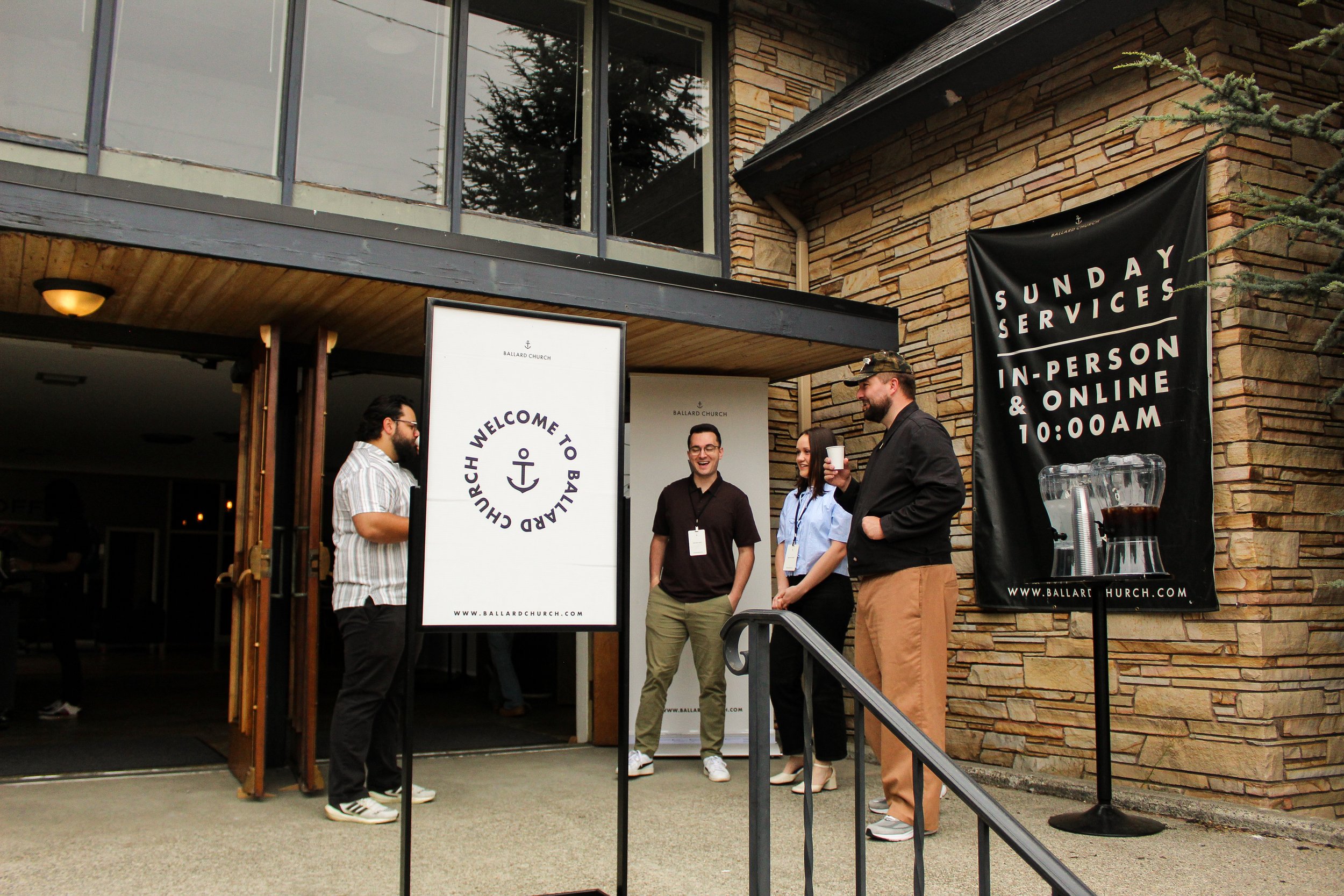 People welcoming visitors outside Ballard Church with Sunday services signage and a beverage station.