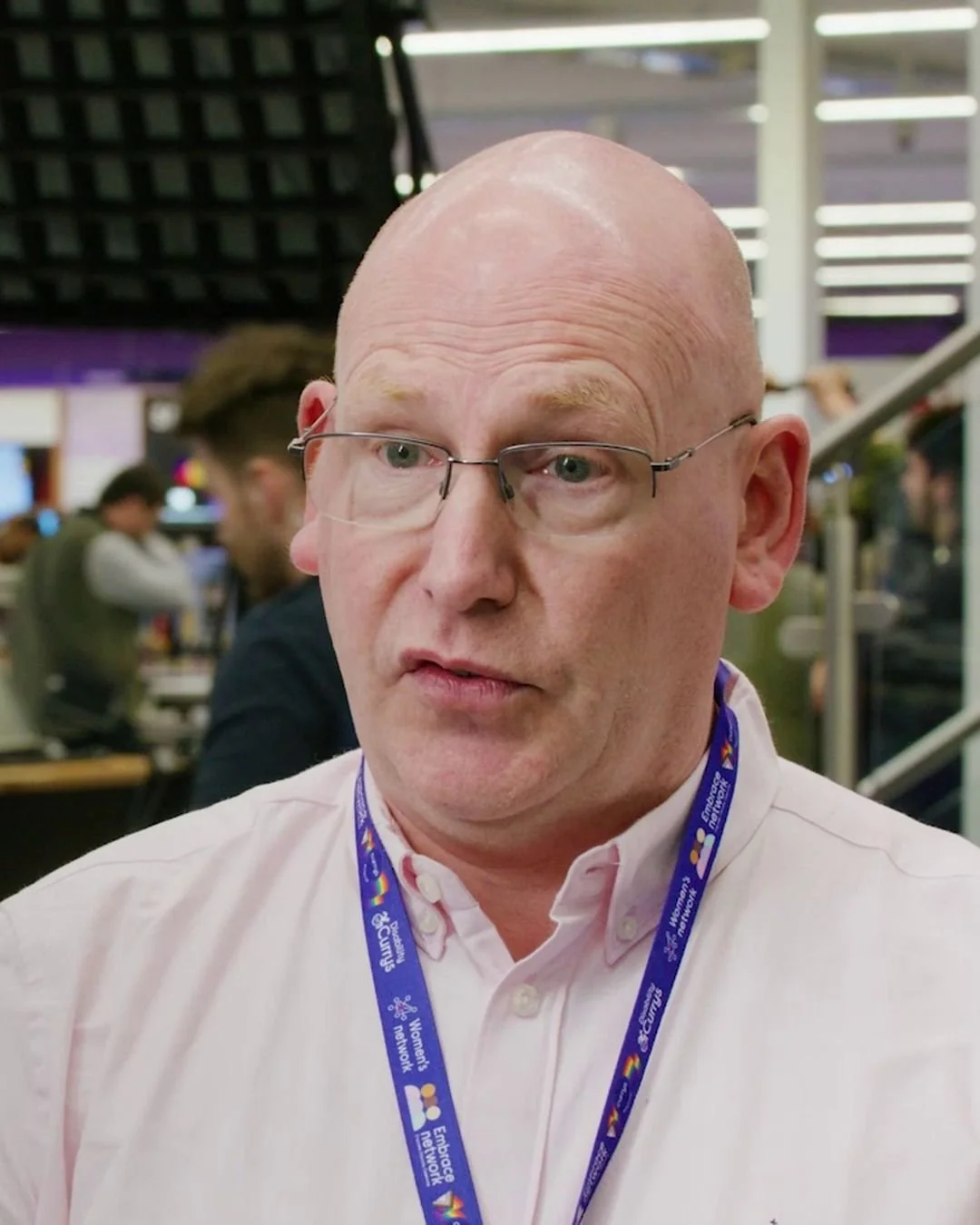 Chris Sadler, a bald man in glasses and a light pink shirt with a Currys colleague networks lanyard, mid-conversation with a thoughtful expression. Behind him, out of focus, is a busy studio set with lighting rigs and people working.