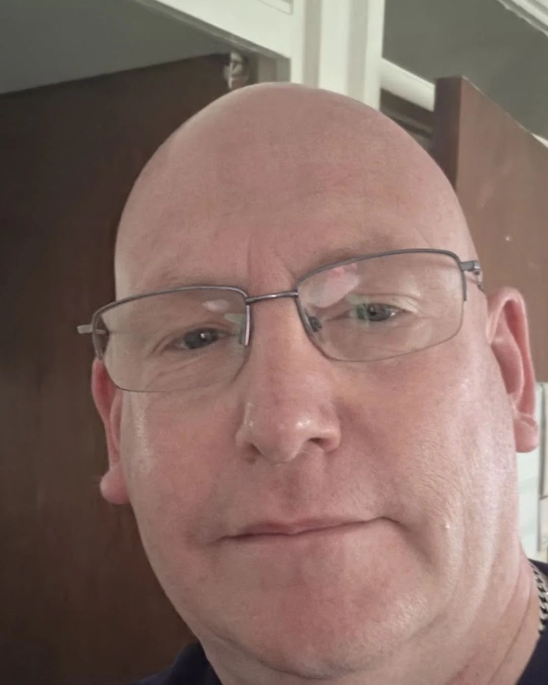 Close-up selfie of Chris Sadler, a bald man with rimless glasses and a navy T-shirt, looking at the camera with a calm, slight smile, indoors in front of a wooden door.