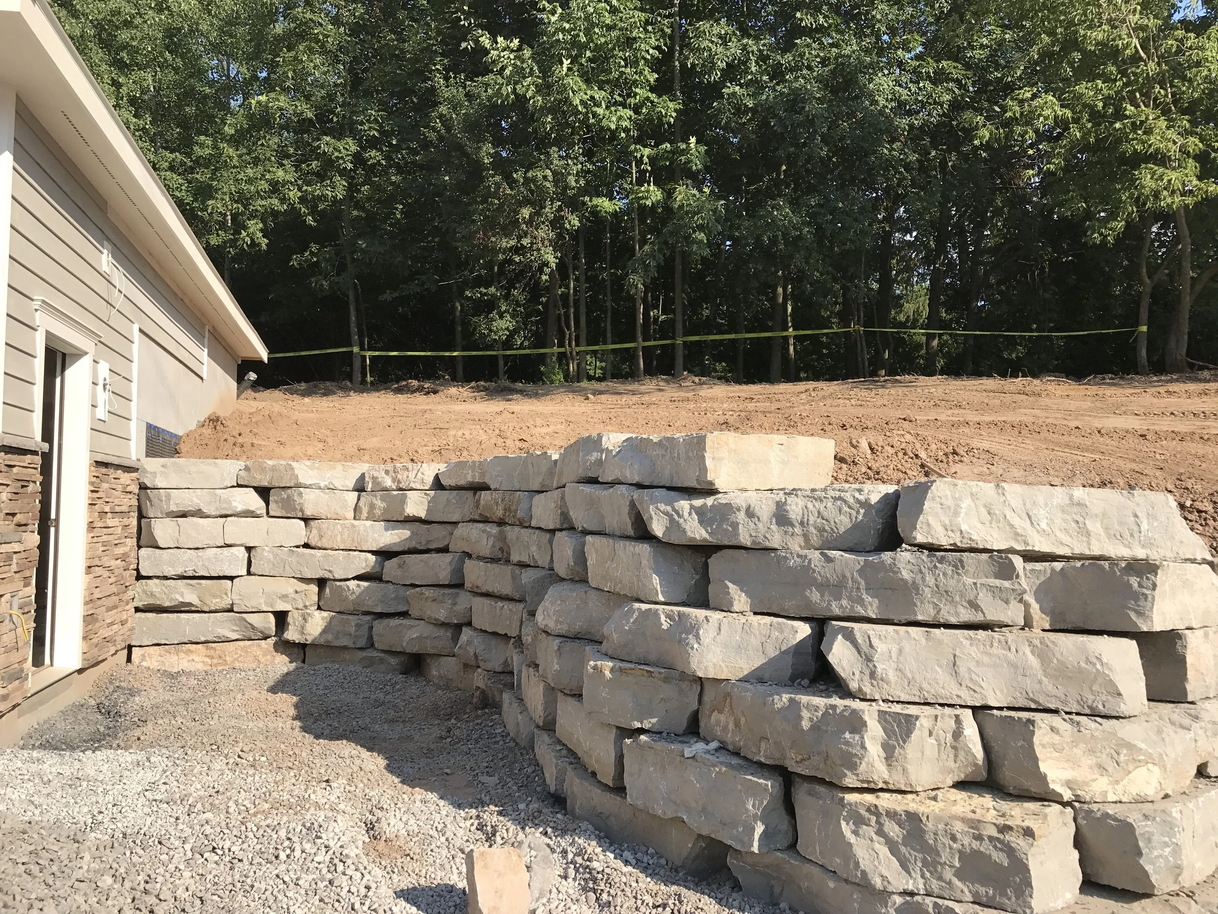 Construction site with a stone retaining wall next to a house with vinyl siding, bare dirt slope in the background, and trees beyond.