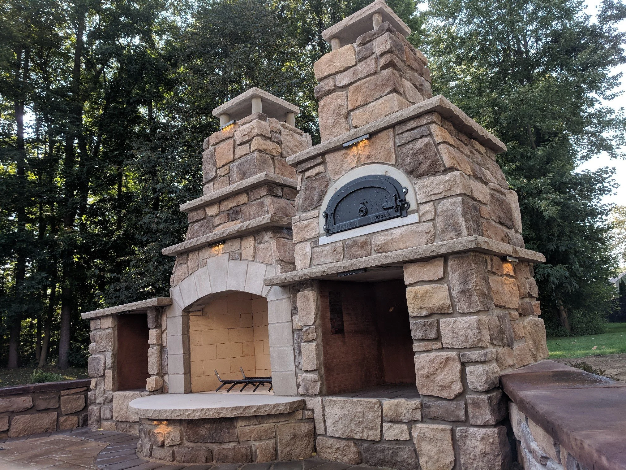 Stone outdoor fireplace with a pizza oven in a backyard with trees.