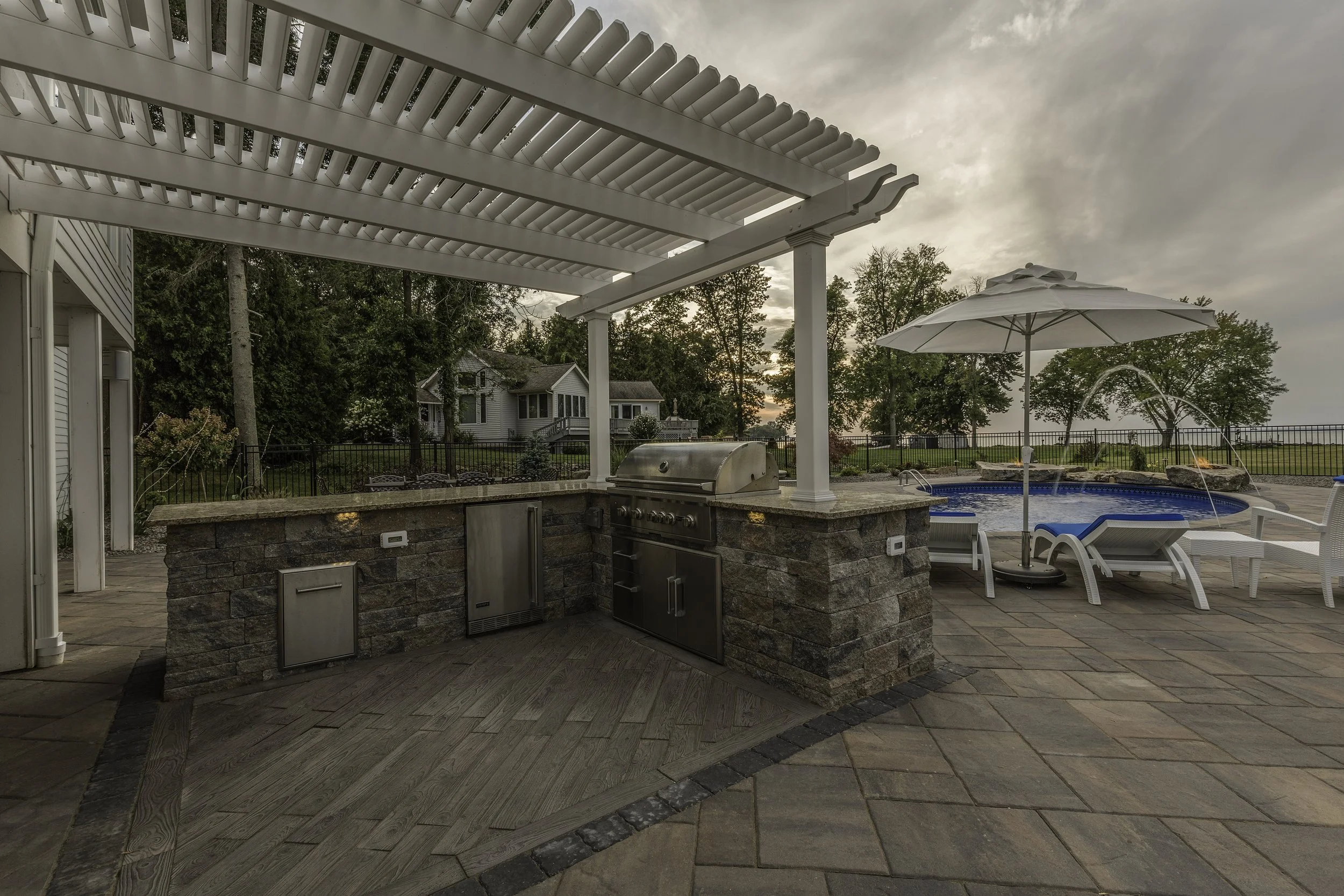 Outdoor patio with built-in barbeque, stone counter, and sunshade, adjacent to a swimming pool with lounge chairs and umbrella, surrounded by trees at sunset.