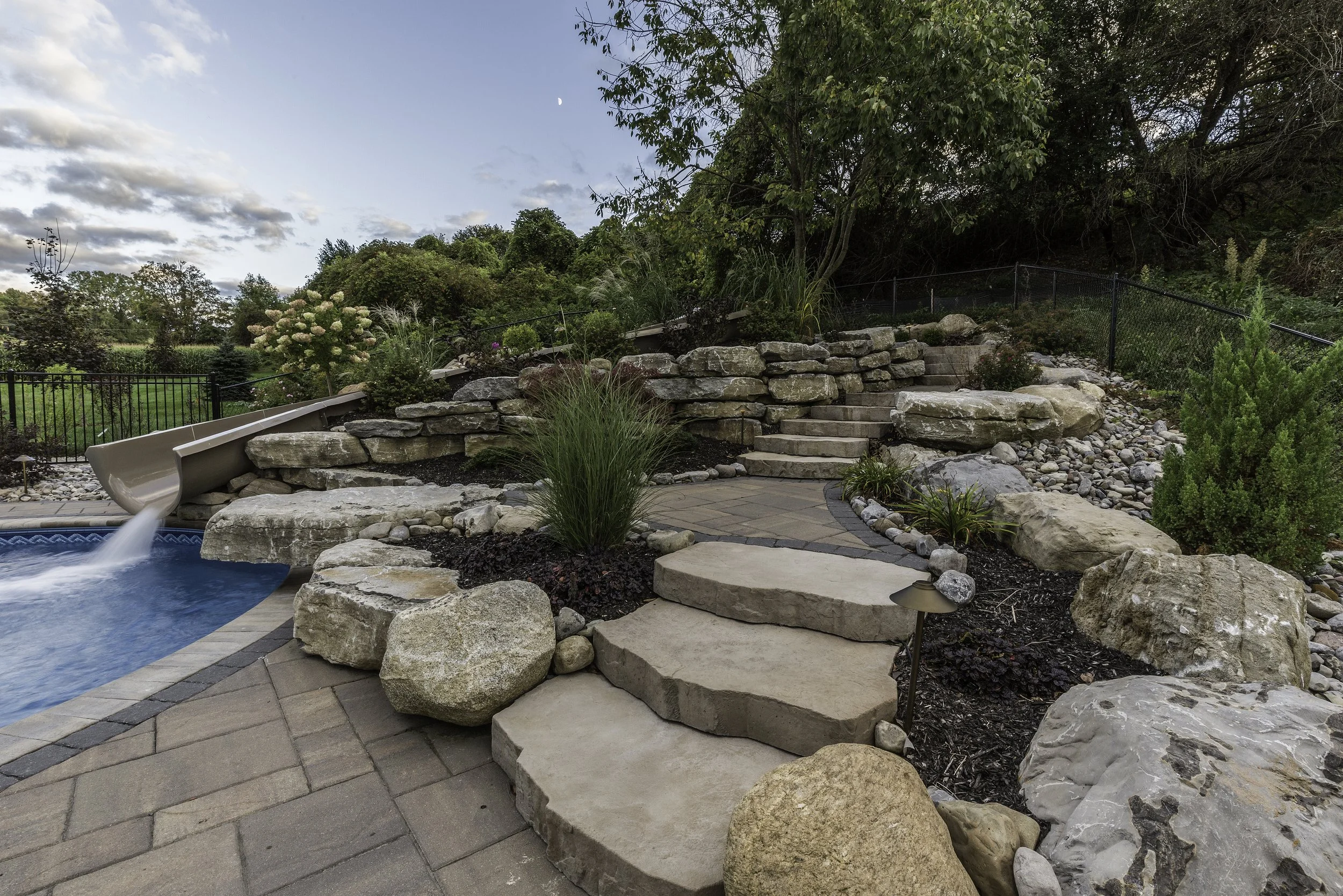 Backyard with swimming pool, waterslide, stone steps, and landscaped garden with plants and trees under blue sky