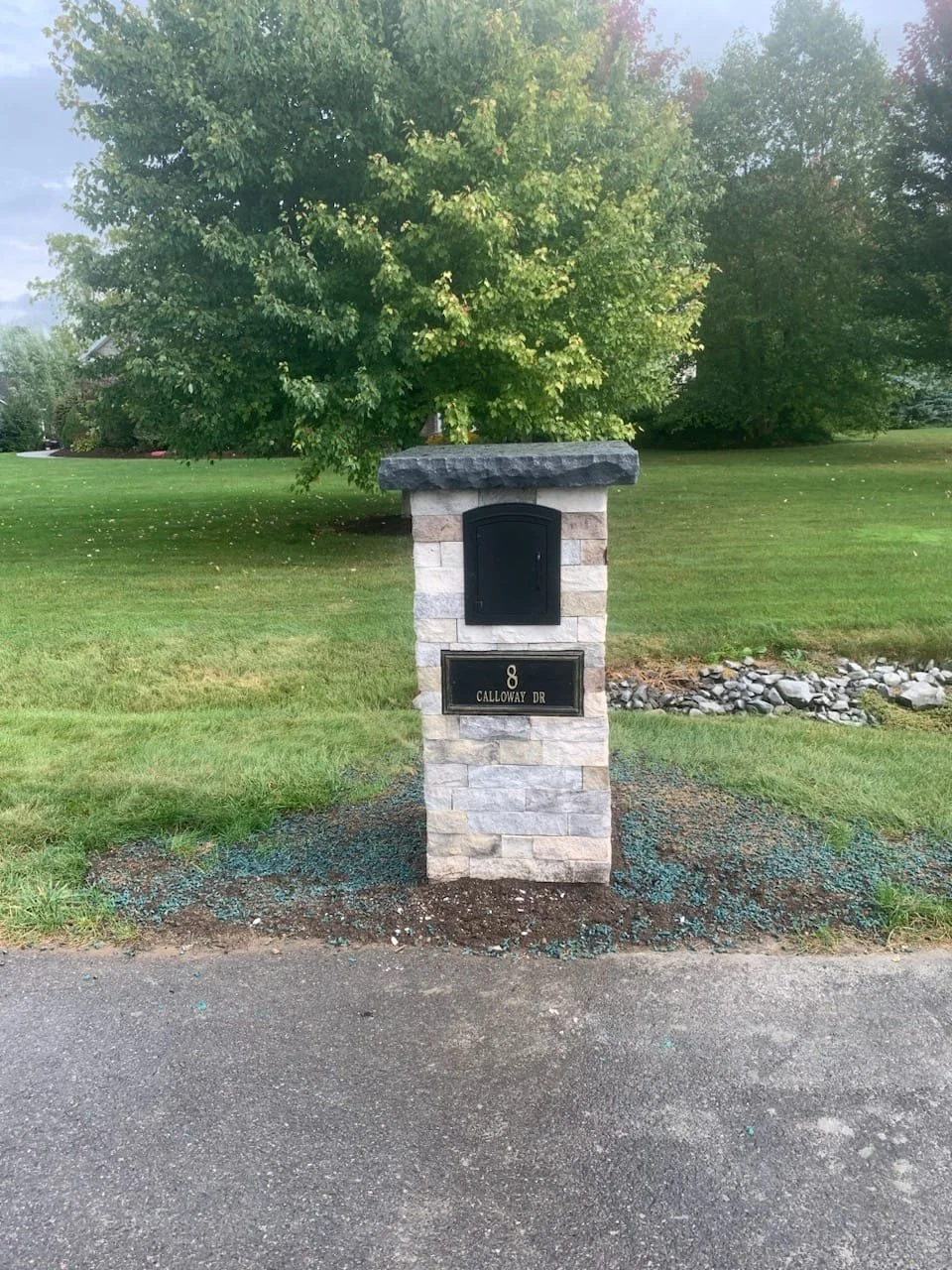 A stone mailbox with a dark metal door and a black house number plaque reading 8, located on Calloway Drive, with a large green tree and a grassy lawn in the background.