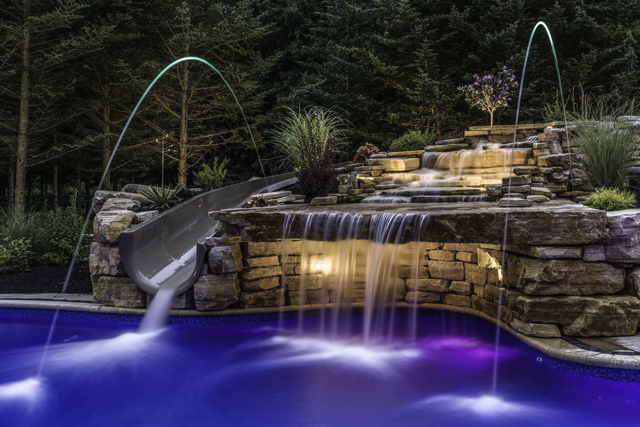 A backyard with a stone waterfall feature and water slides over a blue pool with colorful LED lights.