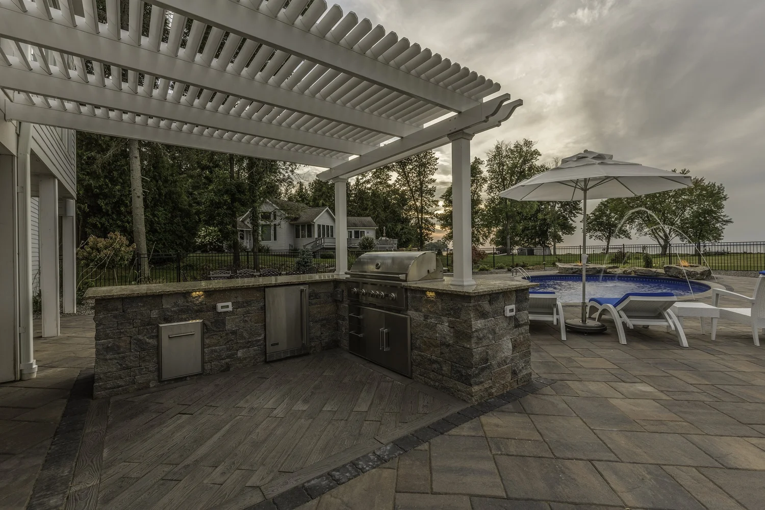 Outdoor kitchen under a white pergola with stone counters near a pool in Central NY